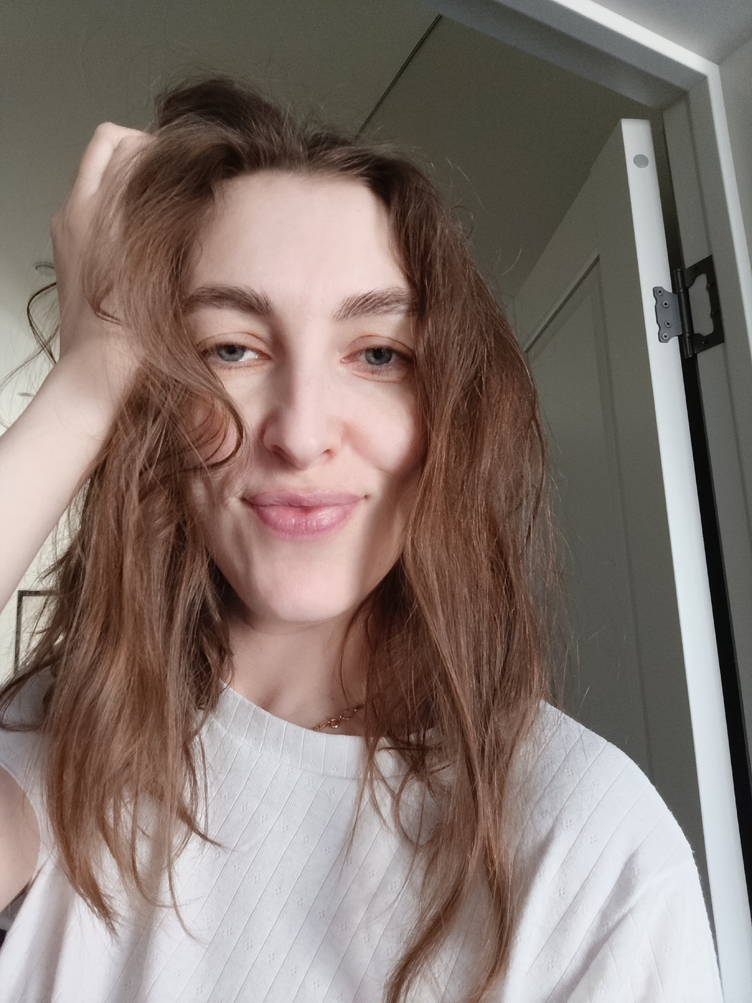 A close-up portrait shows a young woman, reportedly located in Chaikovsky, Russia, with long, wavy brown hair and blue eyes, smiling gently at the camera. Her right hand is raised, with fingers running through her disheveled hair, creating a relaxed and natural pose. Her expression is soft and engaging, with a slight, almost mischievous smile on her lips.
She is wearing a light-colored, possibly white or off-white, short-sleeved t-shirt with a subtle ribbed texture and small, embossed dot patterns. A delicate, gold-toned chain necklace is visible around her neck. Her skin appears fair and her makeup seems minimal, emphasizing a natural look.
The background is simple and domestic, featuring a light-colored wall or ceiling in the upper left. To the right, a white door frame and a slightly ajar white door are visible, with a prominent black hinge on the door. The lighting appears soft and natural, suggesting an indoor setting during daylight hours. No specific weather conditions are discernible. There is no visible text in the image. The overall mood is casual, intimate, and friendly.