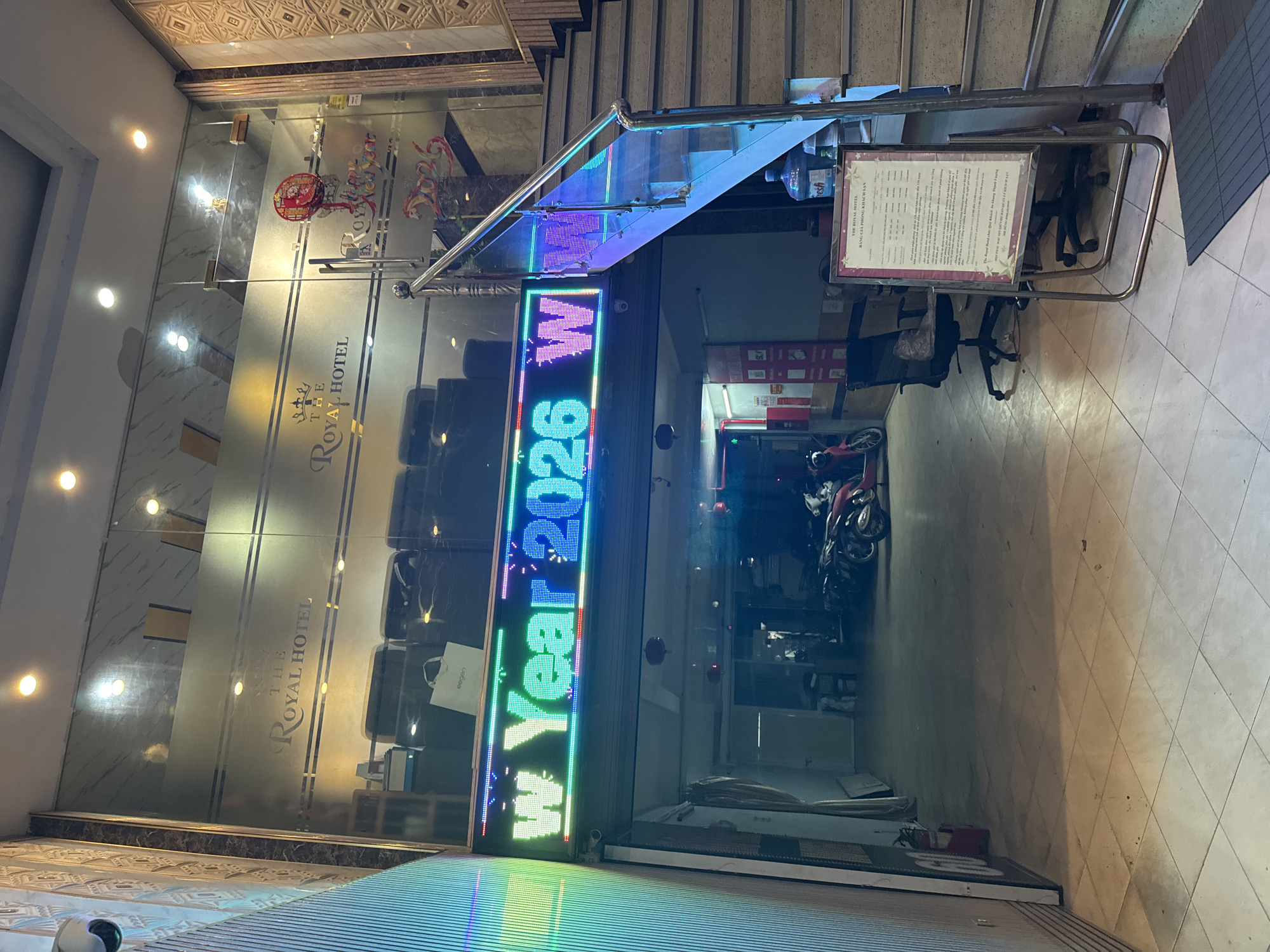 The image shows the entrance to The Royal Hotel in Hanoi, Vietnam. A large, brightly lit LED sign displays "Happy New Year 2026". The hotel's name is also visible etched onto the glass doors. Various objects are present, including a red Chinese lantern, a water bottle, and office chairs. A motorcycle is parked to the right of the entrance. The scene appears to be indoors, possibly in the evening, given the artificial lighting. The overall impression is that of a welcoming hotel entrance, perhaps decorated for a festive occasion. The text visible includes "THE ROYAL HOTEL", "Happy New Year 2026 WW", and "BẢNG GIÁ PHÒNG KHÁCH" (which translates to "Hotel Room Price List" in Vietnamese).