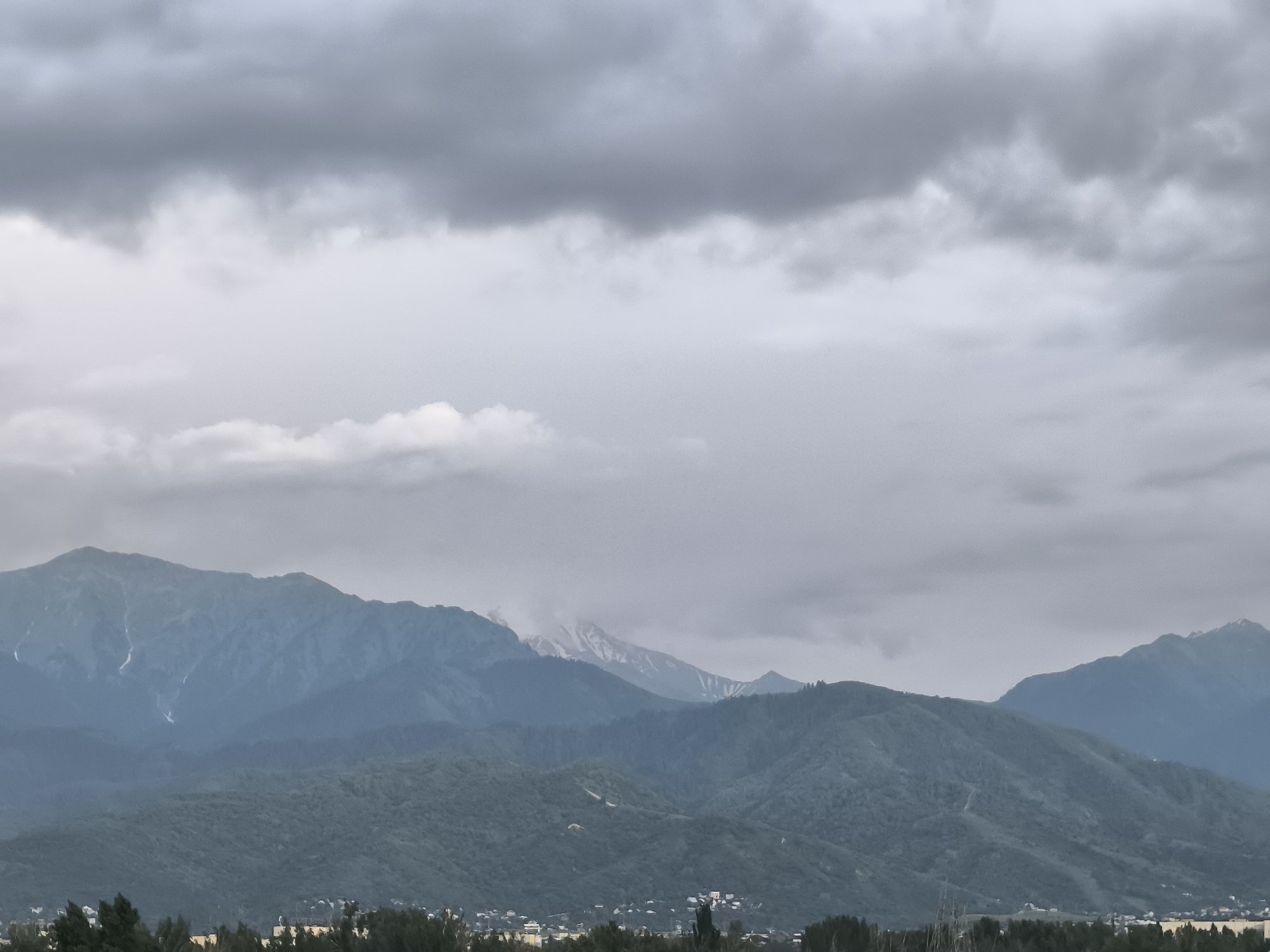 This image captures a majestic mountain range under a cloudy sky, likely taken from Almaty, Kazakhstan. The scene is dominated by the imposing peaks of the mountains, which are a deep blue-green hue, suggesting they are covered in dense vegetation or dark rock. Some of the higher peaks show patches of snow, indicating a considerable altitude.
The sky is filled with a thick layer of gray clouds, with hints of lighter, scattered clouds lower down. This overcast weather creates a subdued, moody atmosphere. The light is diffused, casting a soft glow over the landscape and minimizing harsh shadows. The time of day appears to be late afternoon or early evening, judging by the soft, diffused light and the muted colors.
In the foreground, a line of dark green trees forms a natural border, separating the viewer from the middle ground. Below the main mountain range, there are rolling hills covered in what appears to be dense forest. A small town or village is nestled in the valley between the hills and the main mountains, with scattered buildings and lights visible. The overall impression is one of natural grandeur and a sense of peaceful, rural life at the foot of imposing mountains. There is no visible text in the image.