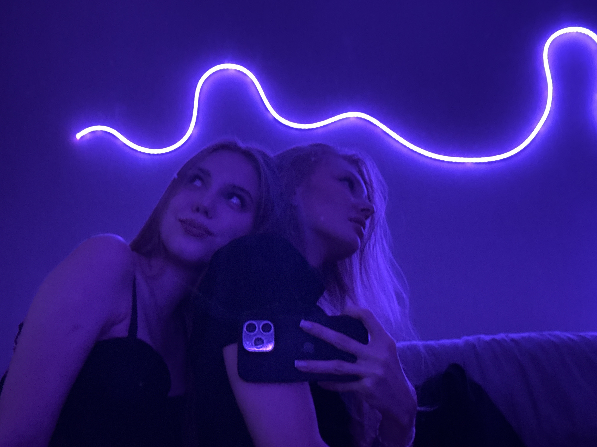 Two young women are in a dimly lit room, with a purple neon light strip forming a wavy line across the upper background. The woman on the left, with blonde hair, is looking upwards and to her left, smiling slightly. She is wearing a black spaghetti strap top. The woman on the right, also blonde, is leaning her head against the other woman's shoulder and holding a smartphone. Her face is mostly obscured, but her eyes are looking towards the right. The phone's camera module is visible, appearing to have three lenses. The environment is generally dark, with purple light dominating the scene. There is no indication of the location being in the city of Perm, Russia.