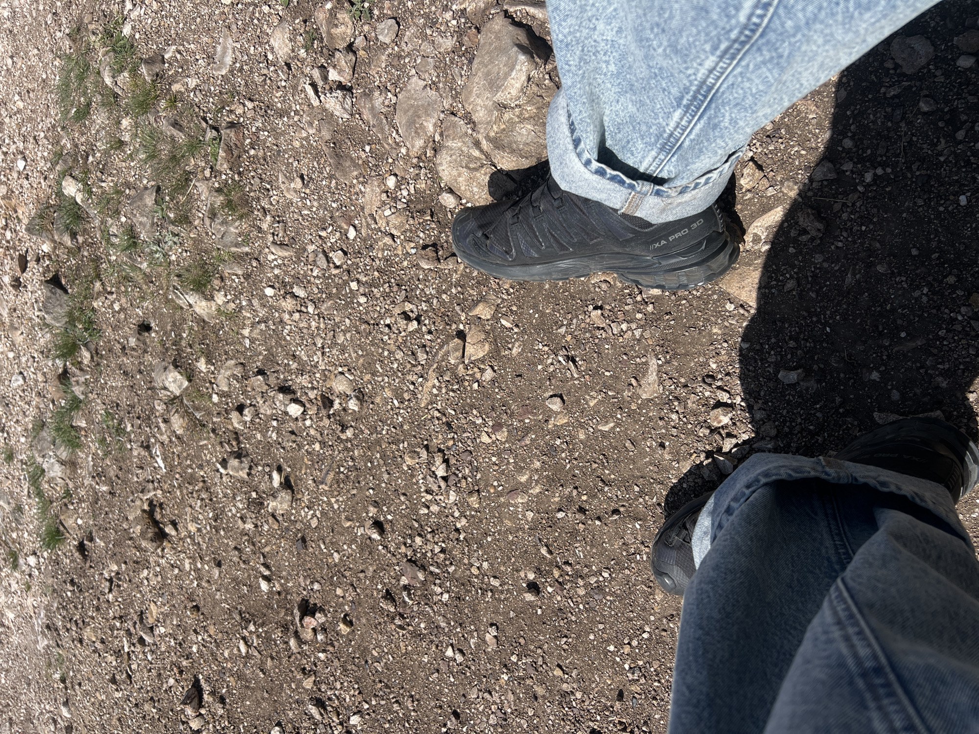 The image shows a gravel path with two feet wearing blue jeans and black Salomon XA Pro 3D shoes. The path appears to be outdoors, with some sparse grass and rocks mixed in with the gravel. The exact location in Krasnoyarsk, Russia, cannot be determined from the image alone.