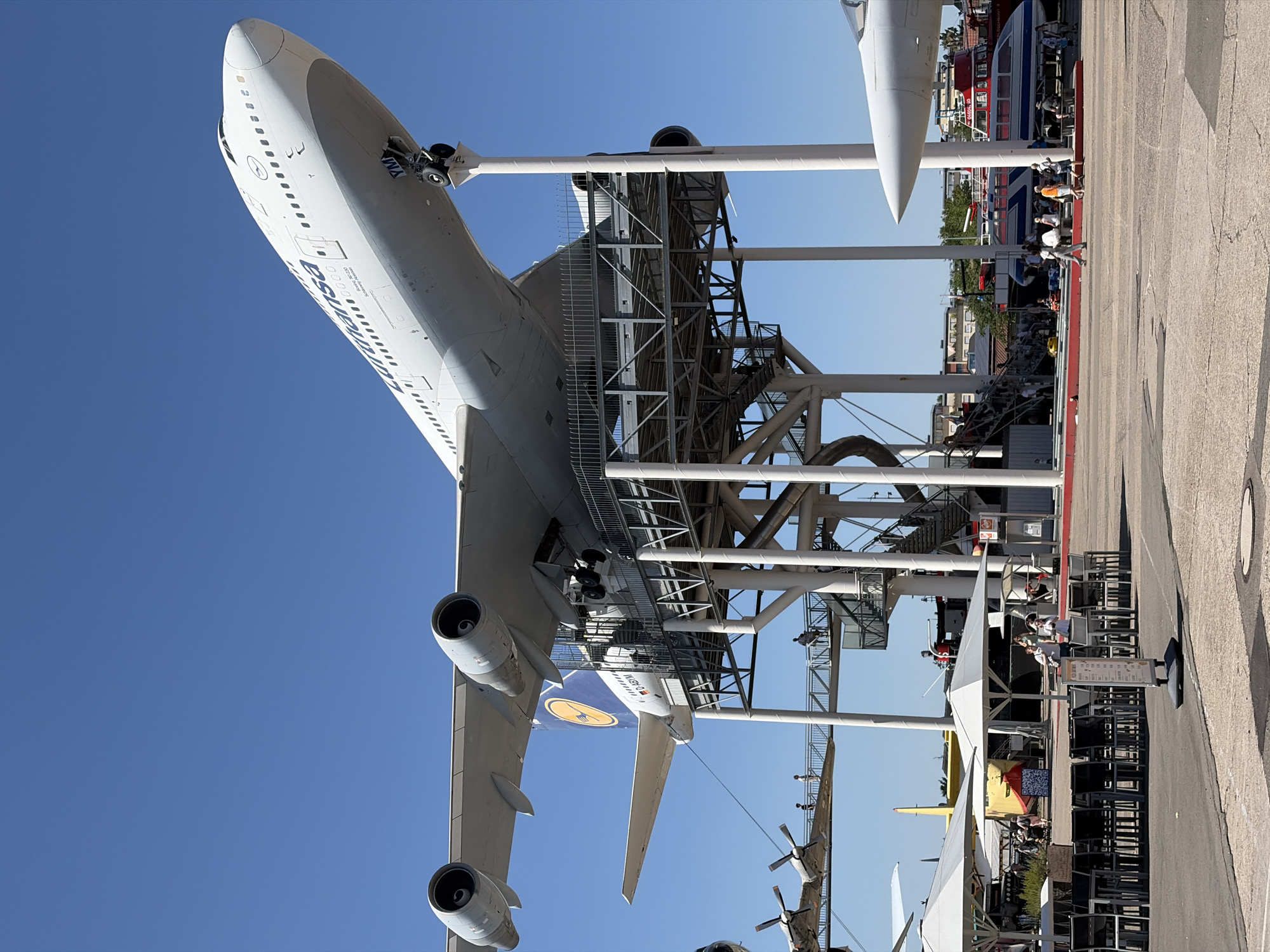 In Speyer, Germany, a decommissioned Lufthansa Boeing 747-230B is displayed on a raised platform, its nose pointing upwards at an angle. The aircraft, registered as D-ABYM, features the airline's blue and yellow livery on its tail fin. Surrounding the aircraft is a multi-level metal structure with stairs and walkways, allowing visitors to access the plane. Several people are visible on the walkways and stairs. In the foreground, there are tables and chairs set up outdoors, with white umbrellas providing shade. Other aircraft and exhibits, including a smaller propeller plane and a white fuselage of another aircraft, are visible in the background. A red and white boat is also present in the harbor area. The sky is clear and blue.
