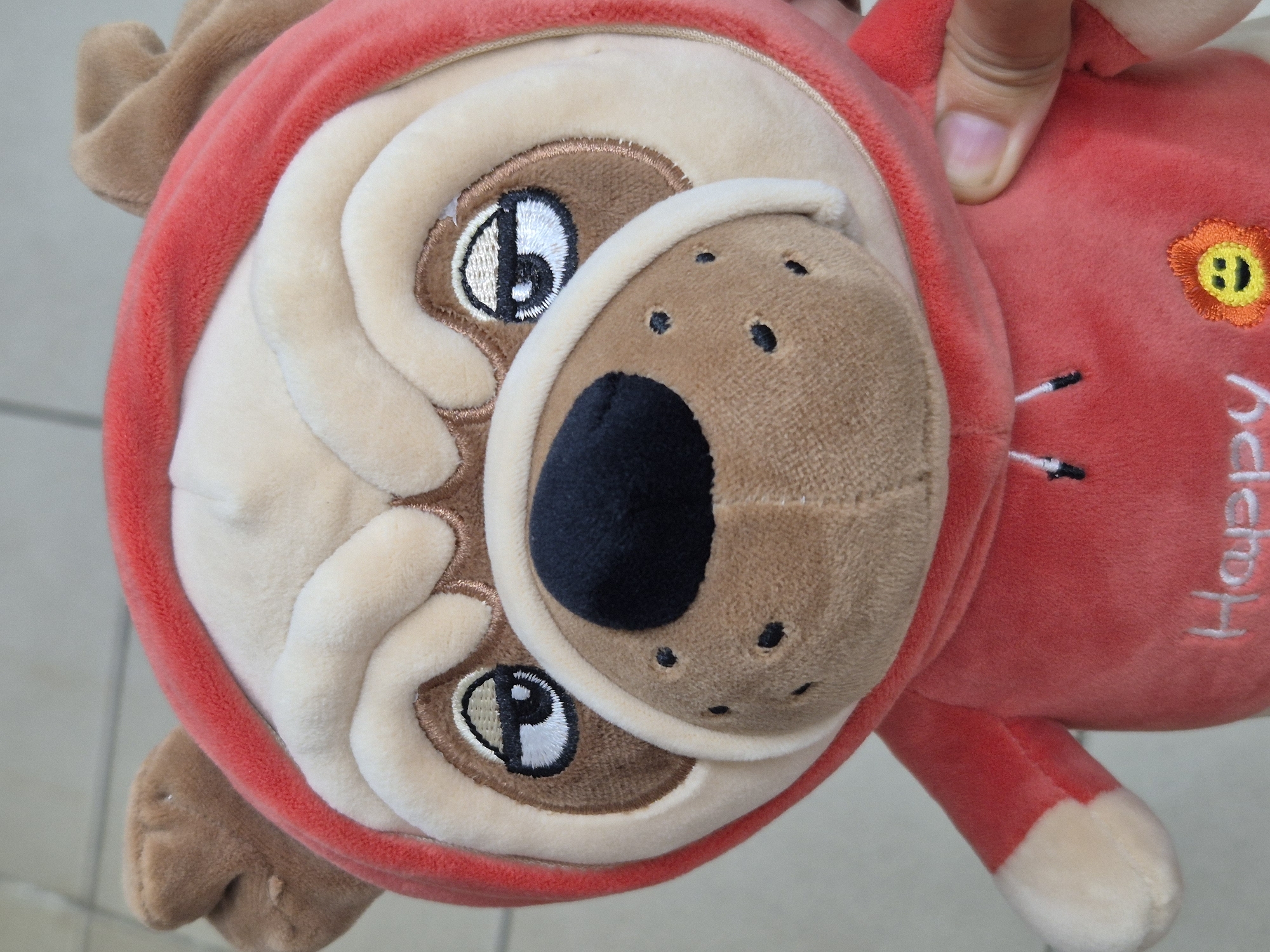 A stuffed toy, resembling a pug, is held by a person's hand. The toy is wearing a red hoodie and has embroidered features including eyes, a nose, and dots on its muzzle. The word "Happy" is also embroidered on the front of the hoodie, along with a small embroidered sun. The background appears to be a tiled floor. This scene is not specifically located in Kokshetau, Kazakhstan.