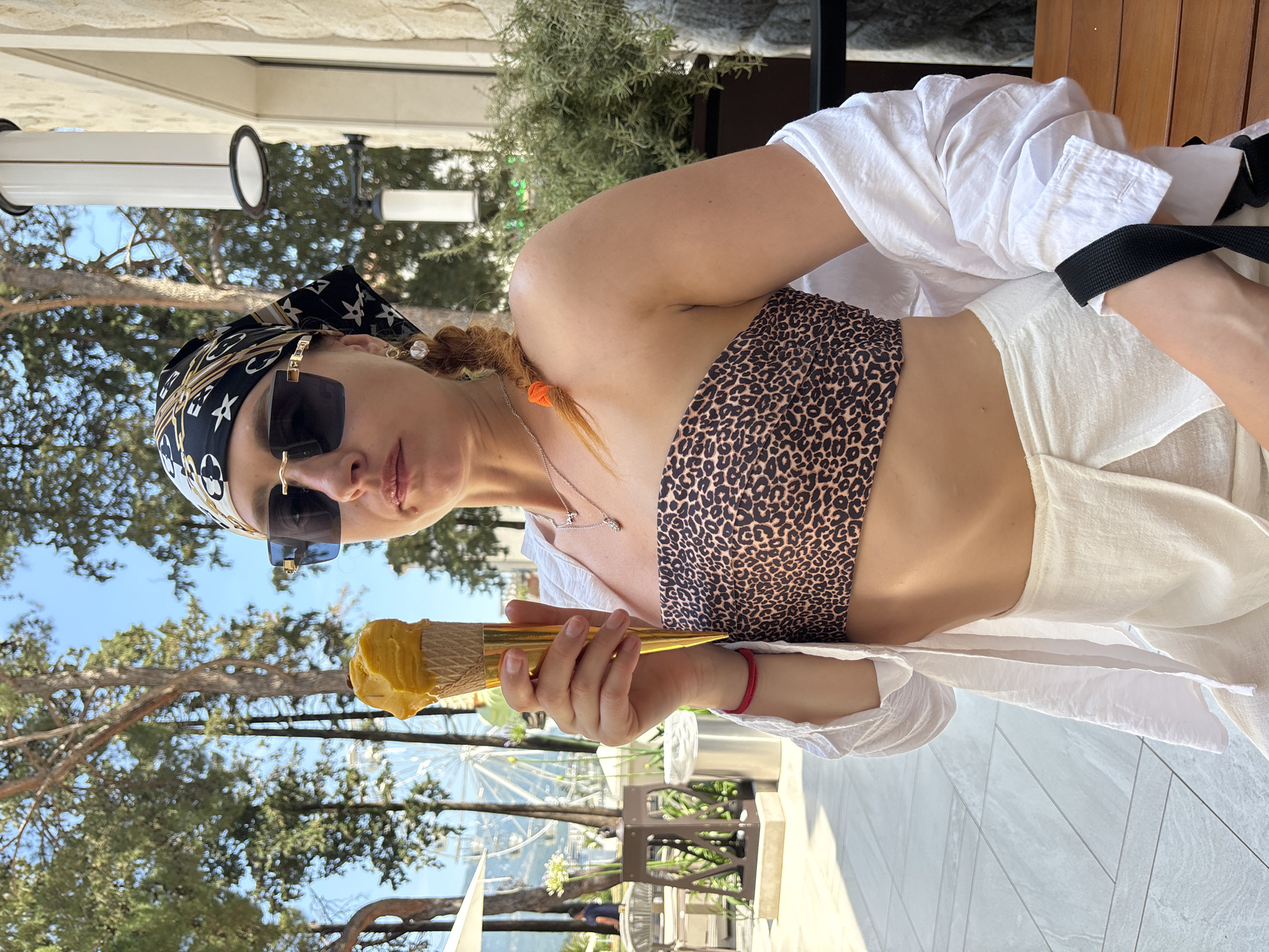 A woman is holding a golden ice cream cone with yellow ice cream while wearing a leopard print bikini top, a white open shirt, white pants, sunglasses, and a black and white patterned headscarf. She has a braid with an orange tie and is wearing a silver necklace and a red bracelet. The background features trees, a Ferris wheel, tables and chairs, and a stone building with lights. The image was taken outdoors in Budva, Montenegro.
