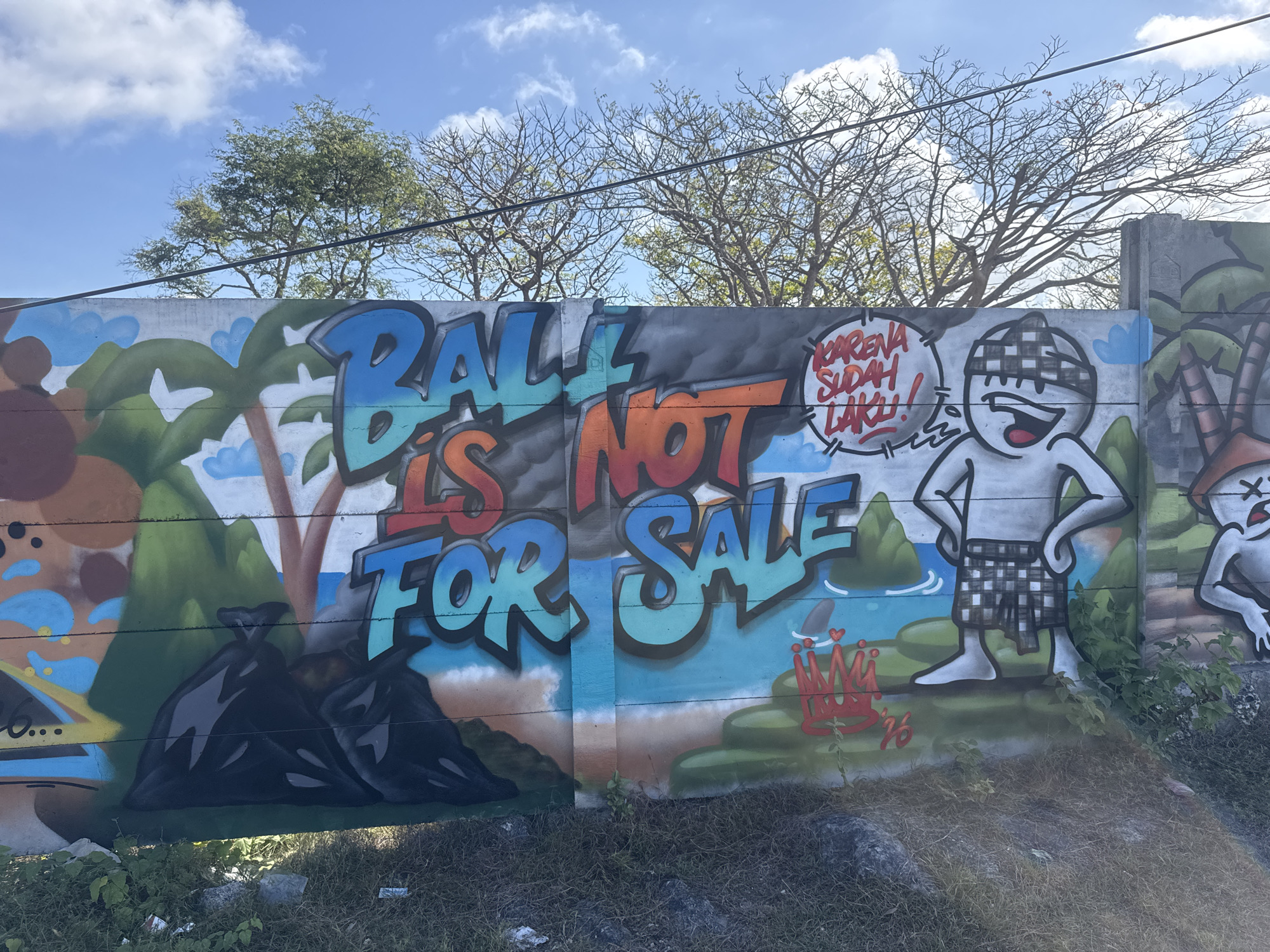 This image shows a colorful graffiti mural on a concrete wall in Kuta Selatan, Indonesia. The mural depicts a tropical landscape with palm trees and a beach scene. In the foreground, there are large letters painted in bright blue and orange that read "BALL IS NOT FOR SALE." To the right of the text, a cartoon character wearing a checkered sarong stands with its hands on its hips. Above the character's head, there is a speech bubble containing red text that says "KARENA SUDAH LAKU!" which translates to "BECAUSE IT'S ALREADY SOLD!" In the lower right corner of the mural, there is a red crown with two hearts above it, and a partial date "76." The wall is situated outdoors, with trees and a blue sky with scattered clouds visible in the background. The ground in front of the wall is covered in dry grass, rocks, and some litter.