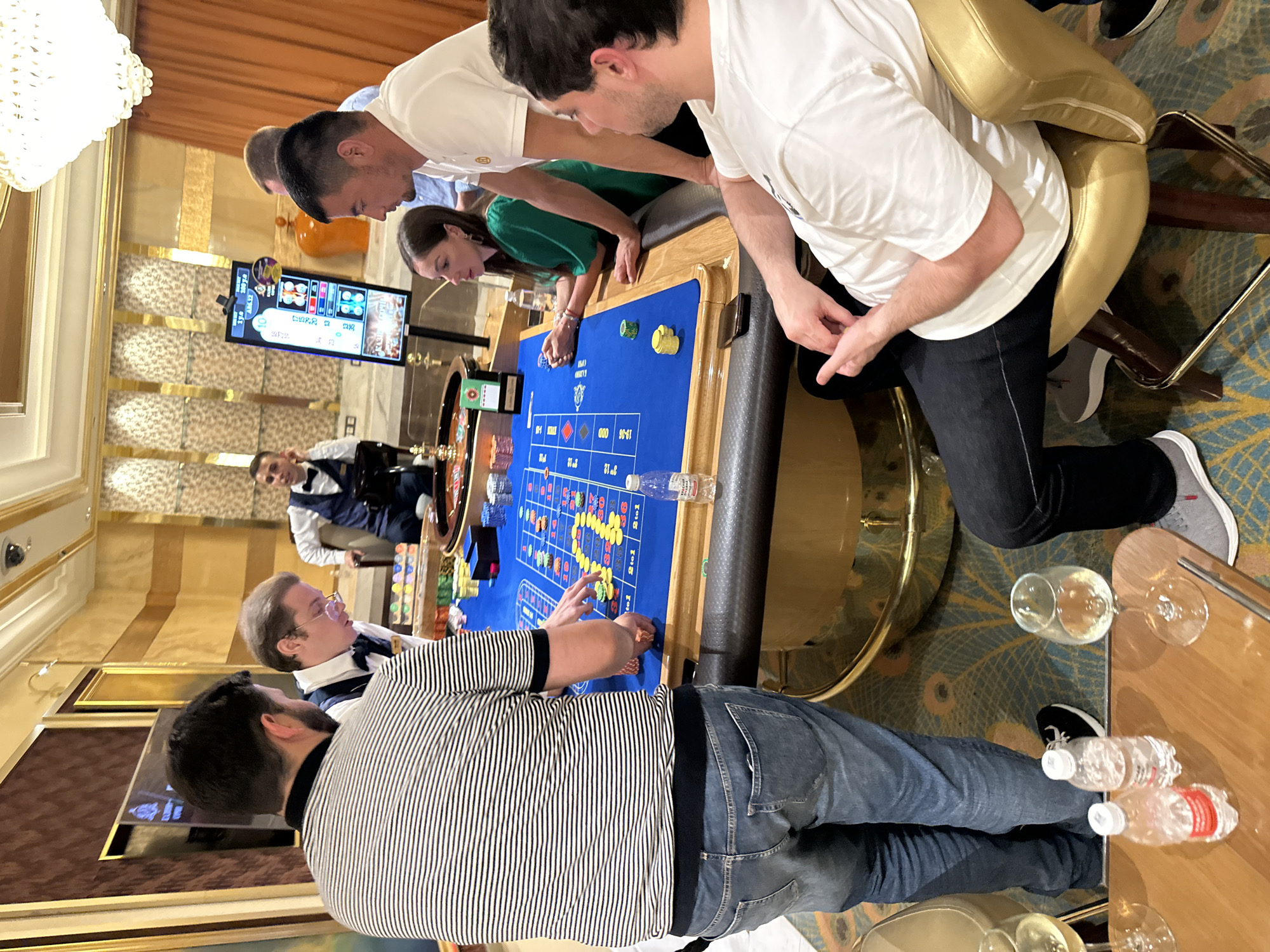 In the city of Estosadok, Russia, a group of people are gathered around a roulette table in a casino. The table is covered in a blue felt surface with betting numbers and patterns. Various colored chips are stacked on the table, indicating bets have been placed. A roulette wheel is partially visible, and a digital display screen shows game information, including numbers and odds.
There are at least six individuals visible in the scene, most of whom are dressed in smart casual attire, with some wearing vests and bowties, suggesting staff or formal attire. One man in the foreground, wearing a striped t-shirt and denim jeans, is looking at the table, with his hand resting near the chips. To his right, another man in a white t-shirt is seated, also observing the game. Across the table, a woman in a green dress is leaning on her hands, watching the roulette wheel. A man in a white shirt and a bow tie appears to be the dealer or croupier, positioned behind the table. Another man in a dark vest and bow tie is seated further back, seemingly in a supervising role. A man in a white shirt and bow tie is also visible in the background, looking towards the digital display.
The environment appears to be an upscale casino interior with ornate decorations, including a large chandelier, patterned walls, and golden accents. The lighting is bright, illuminating the gaming area. On a wooden table in the lower left corner of the image, two bottles of water and a wine glass are present, suggesting that drinks are being served. The overall scene depicts a lively gambling atmosphere in a casino setting.