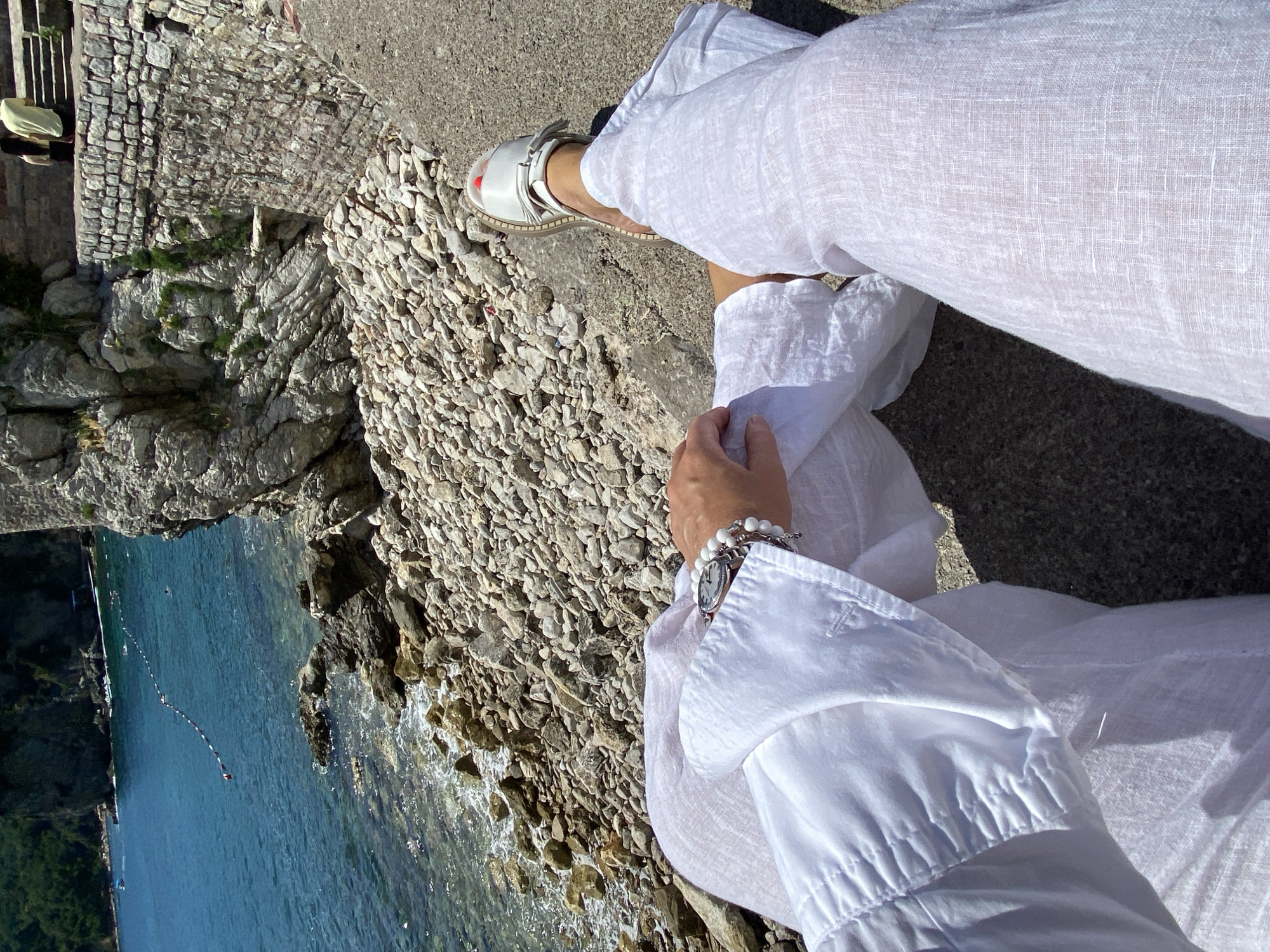 The image displays a first-person perspective view of a person seated on a concrete ledge, overlooking a rocky coastline and the Adriatic Sea in Budva, Montenegro. The individual is wearing loose-fitting white linen trousers and a matching long-sleeved shirt. The left arm, adorned with a silver-toned watch featuring a white face and multiple white beaded and metallic bracelets, rests on the lap. The right foot, visible in the foreground, is clad in a white open-toed sandal, with red nail polish distinctly visible on the toes.
The immediate foreground consists of the concrete surface where the person is seated. Below the ledge, a narrow pebble and rock beach extends to the clear, turquoise water. The sea transitions to a deeper blue further out, with a white buoy line marked with red floats stretching across a section of the water. Two individuals are discernible swimming in the designated area. The background features steep, rugged rocky cliffs rising from the water on the right, partially covered with sparse green vegetation. Above these cliffs, a substantial historical stone wall, constructed from block masonry, is visible, typical of coastal fortifications in the region. Two individuals are seated on a higher stone ledge or steps near this wall. To the left, a darker, heavily vegetated hillside or cliff extends into the sea, with a small boat visible in the distant water. The scene is illuminated by bright sunlight, casting distinct shadows.