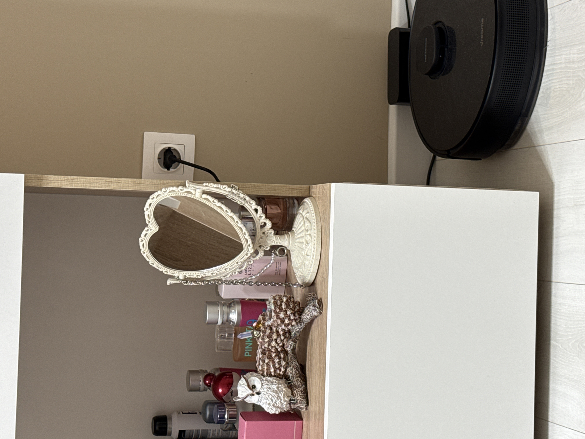 The image shows a shelf with various items on it. There is a heart-shaped mirror, several bottles of various sizes and colors, an owl figurine, and a brown decorative object. A power outlet is visible on the wall above the shelf, and a black robot vacuum cleaner is beside the cabinet. The location appears to be indoors in an unknown city.