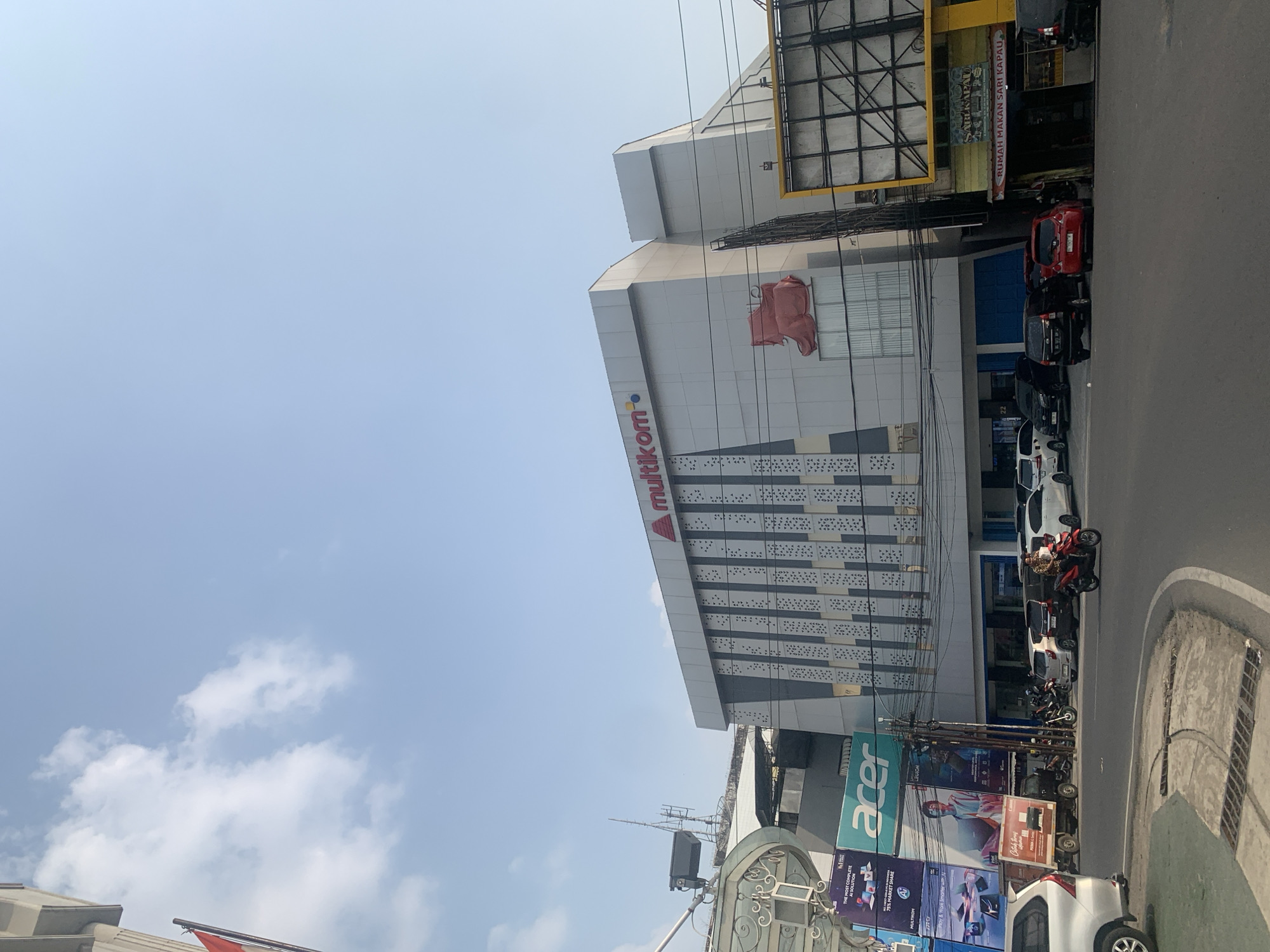 The image shows a street scene in Palembang, Indonesia, with a modern multi-story building labeled "multikom" dominating the background. The building features a grey facade with patterned decorative elements and a large red logo. To the left of the main building, there are advertisements for Acer and other electronic products. A red Indonesian flag is visible flying from a pole on the left side of the frame. In the foreground, a street with several cars and a motorcycle is depicted. A man on a red motorcycle is in motion on the right side of the street. The sky is clear with scattered clouds. The overall scene suggests a commercial area in the city.