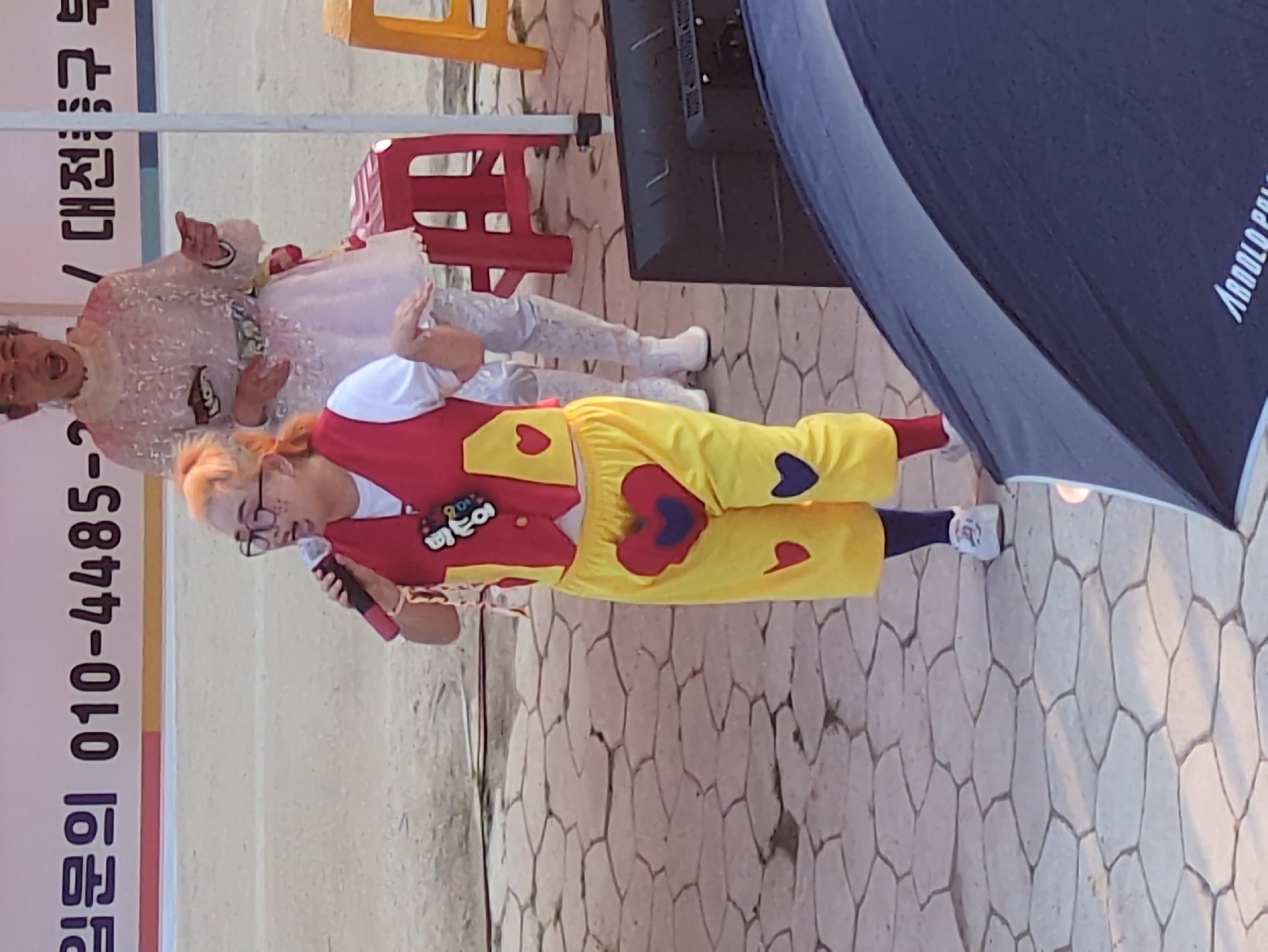 The image shows a man dressed in clown attire, including a red vest with a heart motif and yellow pants also with heart motifs. He is holding a microphone. Another person, possibly a performer, is slightly blurred in the background, wearing a white outfit. They are on a paved surface in an outdoor setting. A banner with Korean writing and phone numbers is visible on the left. The location may be in the city of Daejeon, South Korea, given the Korean text on the banner. There are red and yellow chairs in the background and what appears to be a speaker system. The right side of the image has a large black umbrella.