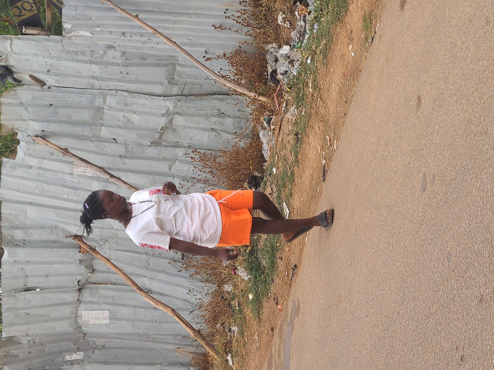 A woman is walking on a paved road in what appears to be a marginal area. She is wearing a white shirt, orange shorts, and sandals. Her hair is pulled back into a ponytail. A corrugated metal fence runs along one side of the road, supported by wooden posts. Debris and vegetation are scattered between the fence and the road. The location is possibly in Jos, Nigeria, based on likely environmental and architectural contexts.