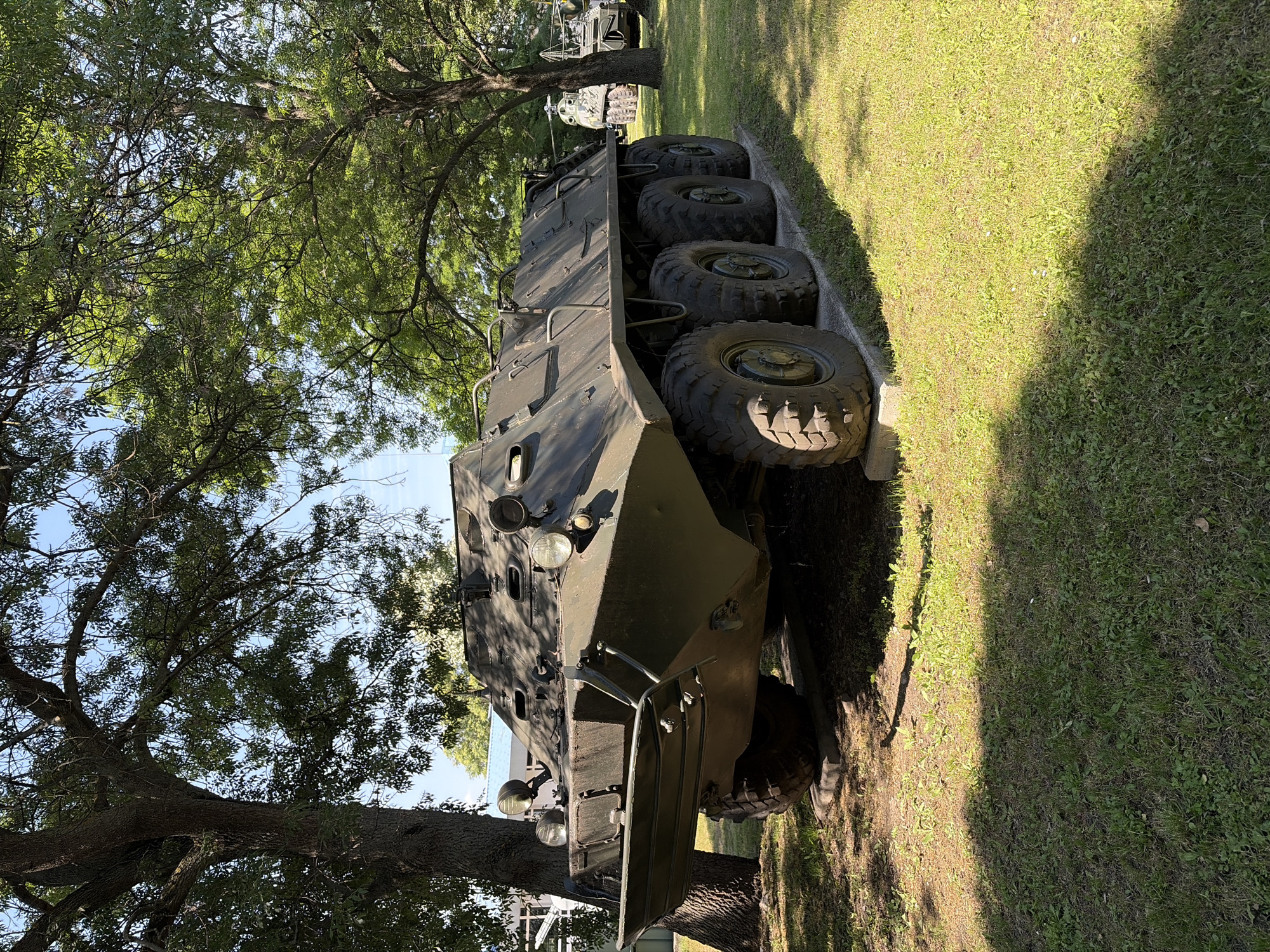 An olive-drab, eight-wheeled armored personnel carrier, likely a BTR-60, is parked on a paved surface in a grassy area. The vehicle is angled towards the left of the frame, with its front facing left. Large trees with dense green foliage provide shade over the vehicle and the surrounding grass. The sky above is clear and blue. In the background, another military vehicle, possibly a helicopter on a trailer, is visible beneath more trees. The setting appears to be an outdoor display or museum, with the BTR resting on concrete blocks. The image was taken in Sofia, Bulgaria, likely at the National Museum of Military History, which features a collection of armored vehicles in an outdoor exhibition area. There are no people visible in the image.