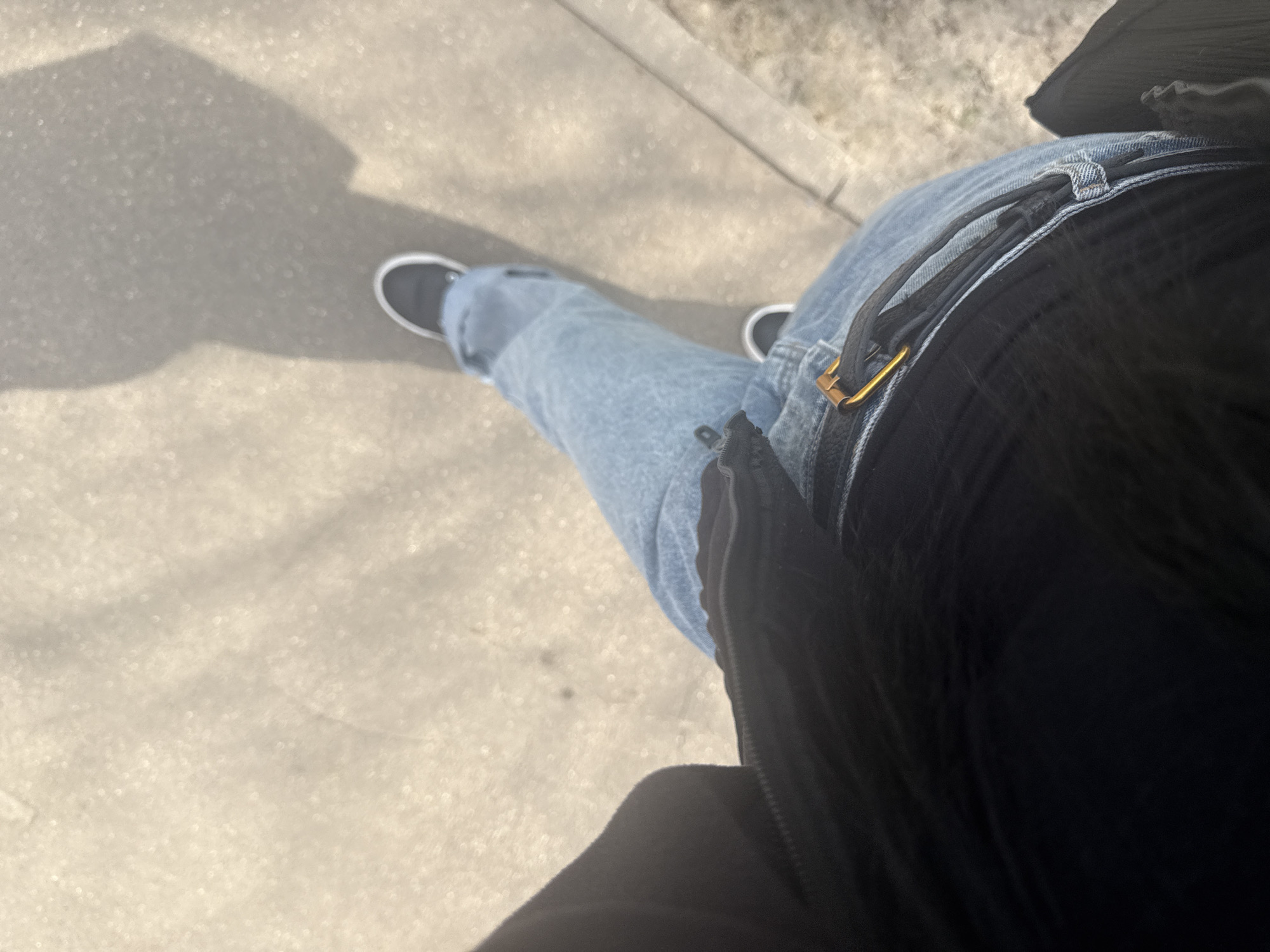 The image presents an overhead view of a person standing on a concrete surface, likely a sidewalk. The person is wearing light blue jeans, a black jacket, and black sneakers. Shadows cast on the concrete indicate the presence of sunlight and possibly trees or other objects creating the shadows. No specific details in the image directly indicate the location is Mytishchi, Russia.