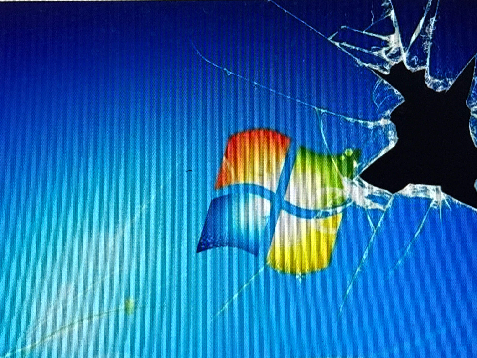 The image is a close-up, angled photograph of a computer screen displaying a cracked screen wallpaper. The wallpaper shows the Windows 7 logo, a multi-colored flag-like shape, against a gradient blue background. Visible cracks spread across the screen, obscuring parts of the logo and the background. The cracks appear as white lines and in some areas create black voids where the screen is broken. The photograph itself seems to have a slight blur and possibly some glare, suggesting it was taken of a physical screen rather than a digital capture of the wallpaper. There are no people, specific city locations, or discernible environmental features other than the computer screen itself.