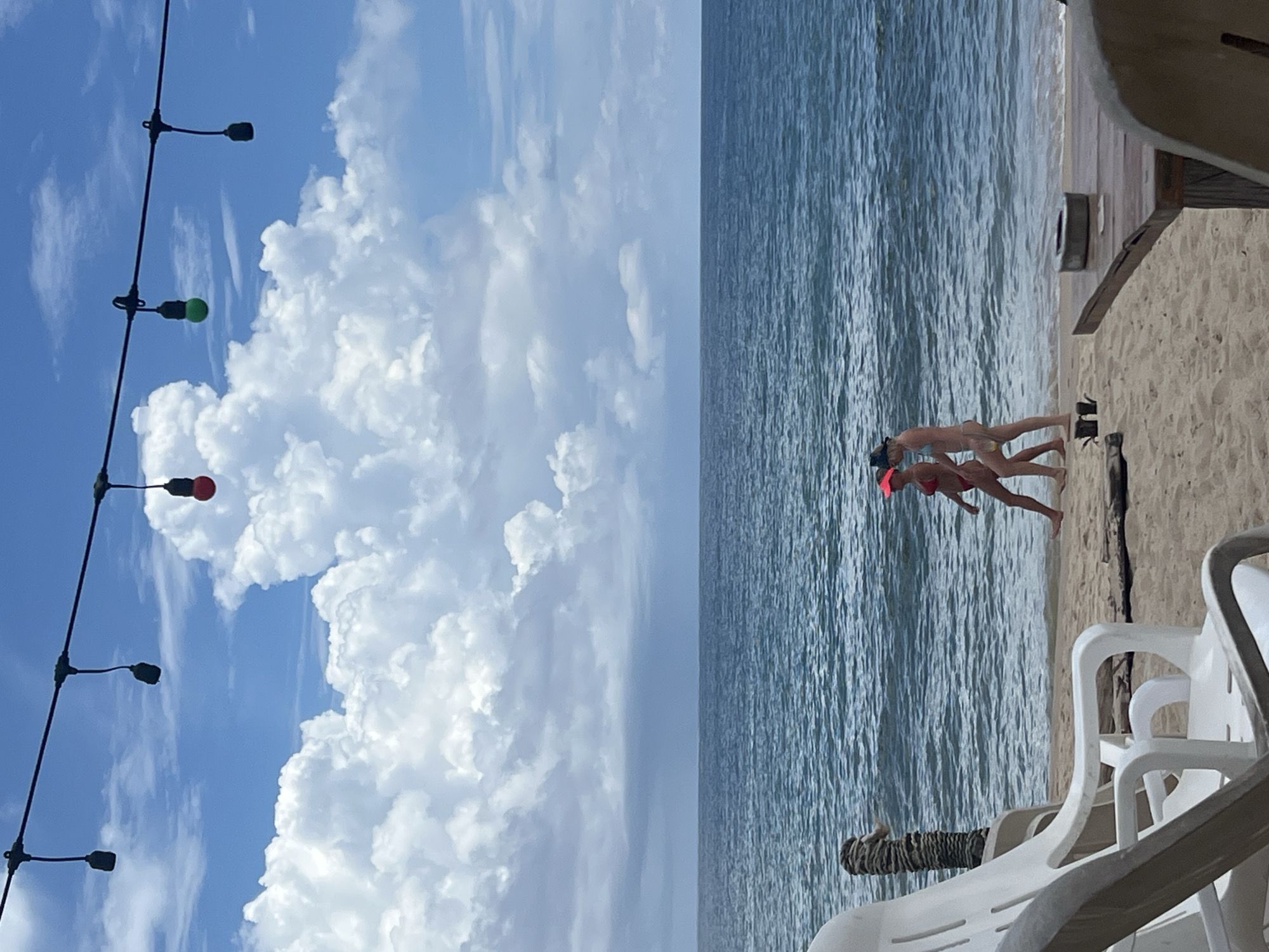 In the city of Choeng Thale, Thailand, two women in bikinis walk along a sandy beach next to the ocean. The woman on the left wears a red bikini and a pink visor. The woman on the right wears a light blue bikini and a dark visor. The ocean is a deep blue with moderate waves. Above them, a string of lights hangs against a sky filled with large, fluffy white clouds. In the foreground, white plastic beach chairs and a wooden table are visible.