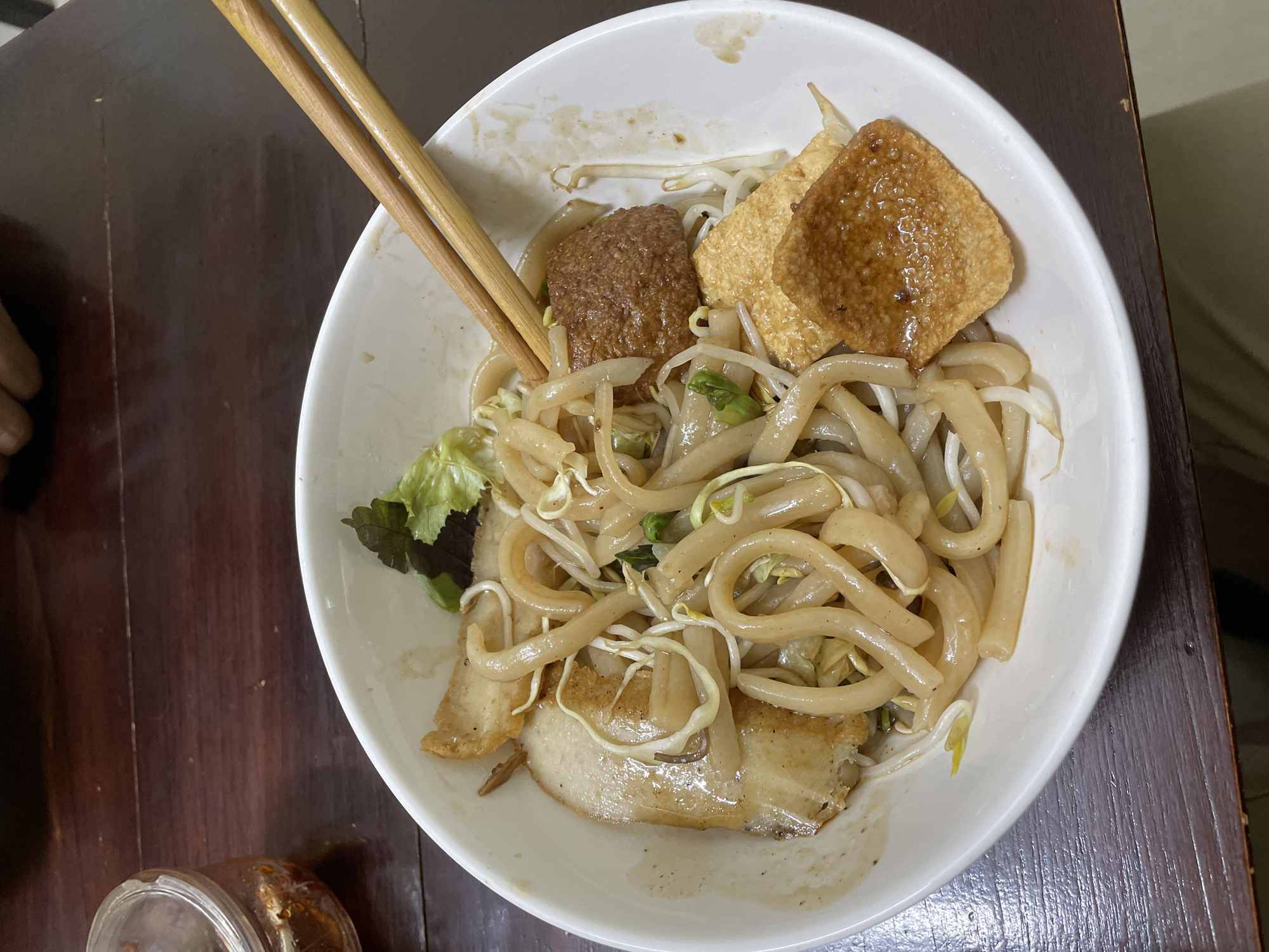 A close-up, overhead shot shows a white bowl of noodles and various toppings resting on a dark wooden surface, likely a table. Chopsticks, made of light-colored wood, are held in the upper left portion of the frame, resting above the bowl. The bowl contains thick, light-colored noodles mixed with bean sprouts and some leafy greens. Among the toppings are two pieces of fried tofu, one square and one rectangular, with a shiny, glazed surface, and a circular patty of ground meat. There are also slices of pork belly with crispy skin visible at the bottom of the bowl. The overall presentation suggests a meal being served or eaten. The lighting appears to be indoor, possibly natural light from a window or artificial light, casting soft shadows and highlighting the textures of the food. The context of Đà Nẵng, Vietnam, places this scene in a Southeast Asian setting, where noodle dishes are a staple. No visible text is present in the image.