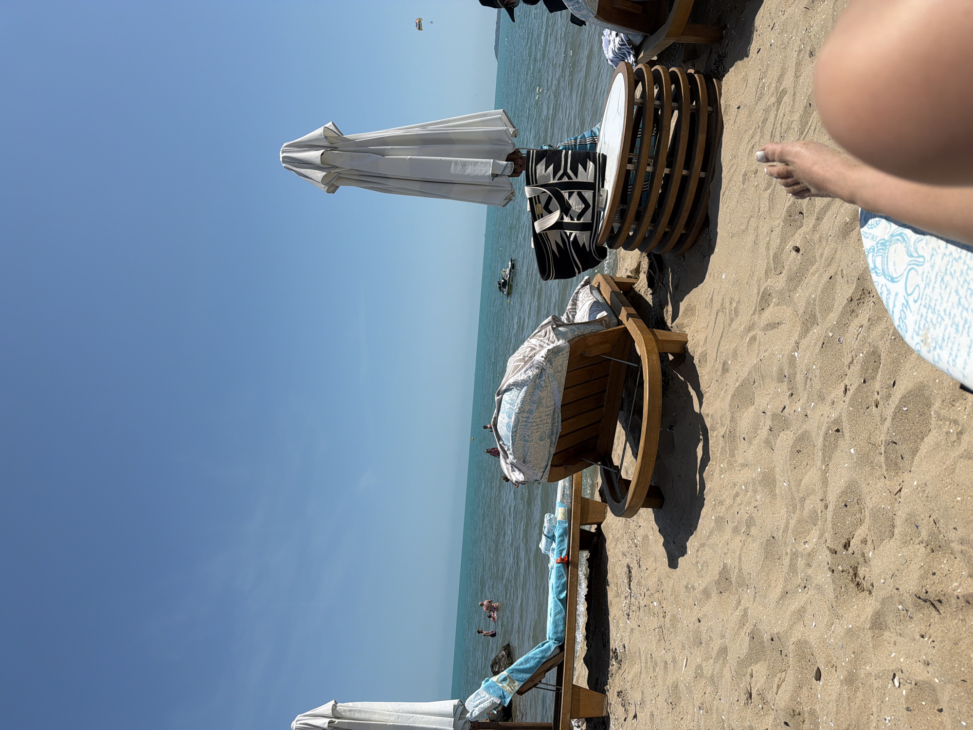 The image depicts an outdoor beach scene on a clear, sunny day in Unknown, Unknown. The perspective is from a low vantage point, presumably from a person reclining in a beach chair. The bottom right of the frame shows a bare, tan right leg and foot with light-colored toenail polish, resting on a blue and white patterned surface, likely a cushion or towel.
The immediate foreground consists of light brown sand, showing various small shells, pebbles, and indistinct footprints. In the midground, two wooden beach lounge chairs are visible. The chair on the left is adorned with a blue patterned towel and positioned closer to the water. The chair in the center-right is partially obscured by a large, upright, closed white or light grey beach umbrella. A geometric black and white patterned beach bag with light handles hangs from the pole of this umbrella, resting on a round, multi-layered wooden table with a white top, situated between the two central chairs.
Beyond the beach chairs, the turquoise-blue ocean extends across the middle and background. Several individuals are in the water; a small group of three to four people wade near rocks in the shallow left side, and another person is visible further out on the right. A white jet ski is present in the water on the left, generating a wake. High in the sky on the far right, a yellow and orange parasail is visible, with a person suspended beneath it. The sky above is a uniform, clear blue, devoid of clouds. Shadows from the beach furniture and umbrella are cast onto the sand, indicating strong direct sunlight.