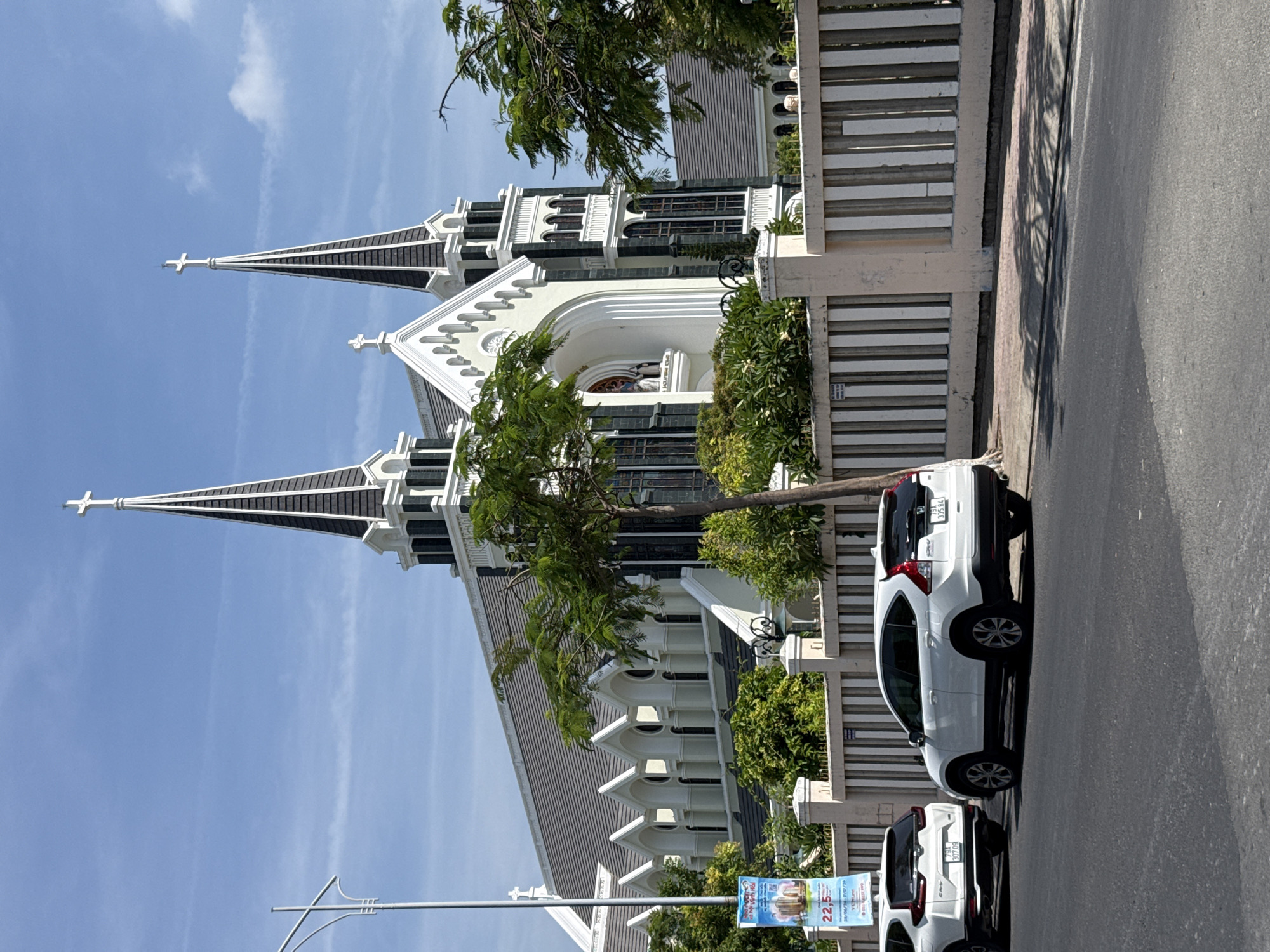 In the city of Bac Nha Trang Ward, Vietnam, a large white church with tall spires stands under a clear blue sky with scattered clouds. The church features dark roofs and trim, with prominent crosses atop its spires. In front of the church, a beige and white striped fence runs along the sidewalk. Two white SUVs are parked on the street in front of the fence. The left SUV is partially obscured by a tree and a banner displaying text and an image. The banner has a price of "22,5" and dates that appear to be in the format DD/MM/YY. The SUV on the right has its license plate visible. Green trees and foliage surround the church and fence, indicating a well-maintained urban environment. A street lamp with a curved arm stands on the left side of the image.