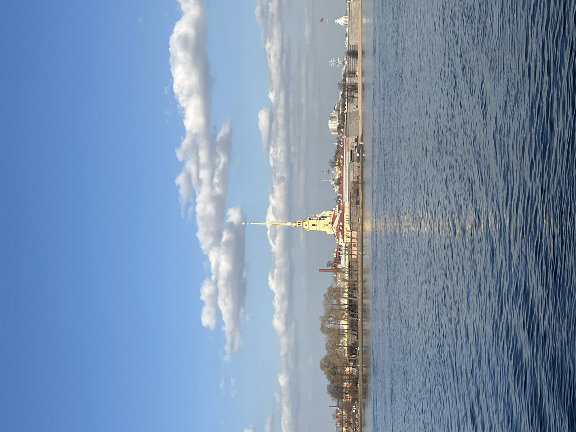 The image captures a panoramic view of the Peter and Paul Fortress and its cathedral in Saint Petersburg, Russia, from across the Neva River. The sky occupies the upper portion, displaying a clear blue expanse with scattered white cumulus clouds. The lower half of the image features the deep blue, rippled surface of the Neva River, reflecting sunlight.
The middle ground is dominated by the Peter and Paul Fortress, situated on what appears to be an island or peninsula. The most prominent structure is the Peter and Paul Cathedral, identifiable by its tall, slender golden spire topped with a cross, rising centrally above the fortress complex. The cathedral features an ochre-yellow facade with white architectural details and a reddish roof. Grey stone fortress walls line the riverbank, with a narrow sandy strip visible at their base in several sections.
To the left of the cathedral, a cluster of bare deciduous trees and various buildings with red rooftops are visible, along with a prominent red industrial chimney. To the right of the cathedral, more fortress structures are present, including a tall mast-like feature resembling a ship's rigging and a white tower-like structure on the far right displaying a red flag, likely the flag of Russia. No identifiable individuals or specific actions are discernible within the frame. The scene is illuminated by natural daylight.