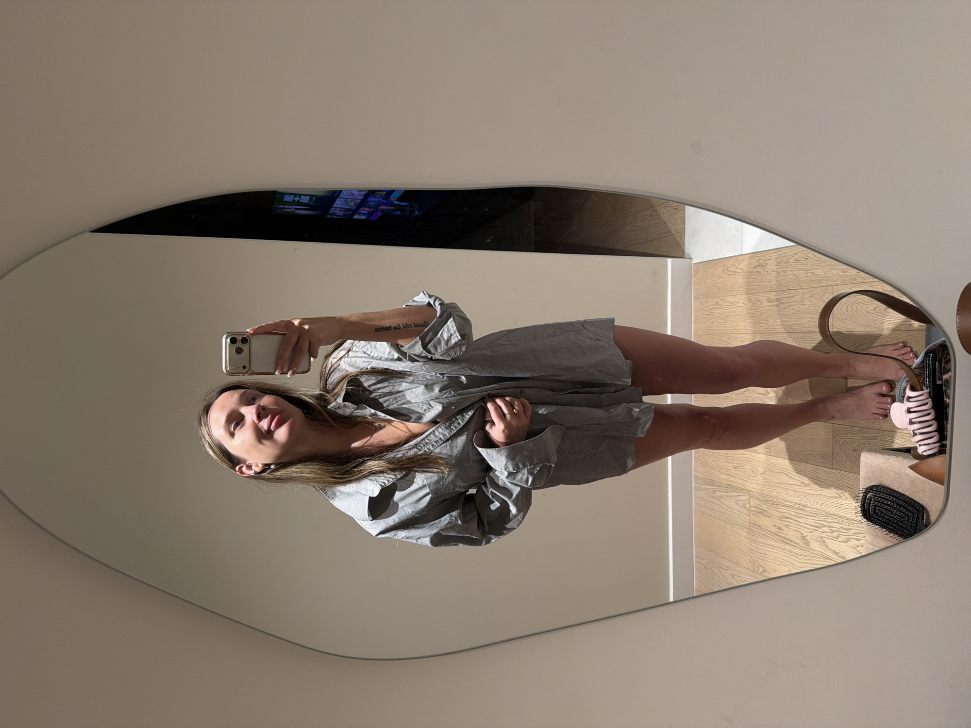 A woman stands in a room, reflected in a large, irregularly shaped mirror. She is wearing an oversized grey button-down shirt and no pants, exposing her legs. She holds a smartphone in her right hand, appearing to take a selfie. Her left hand is placed on her stomach, near the unbuttoned portion of her shirt. The woman has long, brown hair and is wearing earrings and rings. In the reflection, her bare feet are visible on a wooden floor. To her left, a black hairbrush rests on a beige stool, and a brown bag handle is visible. The background of the room is sparsely decorated, with a light-colored wall and a television screen displaying colorful images in the upper right portion of the mirror's reflection. The lighting is soft and ambient.