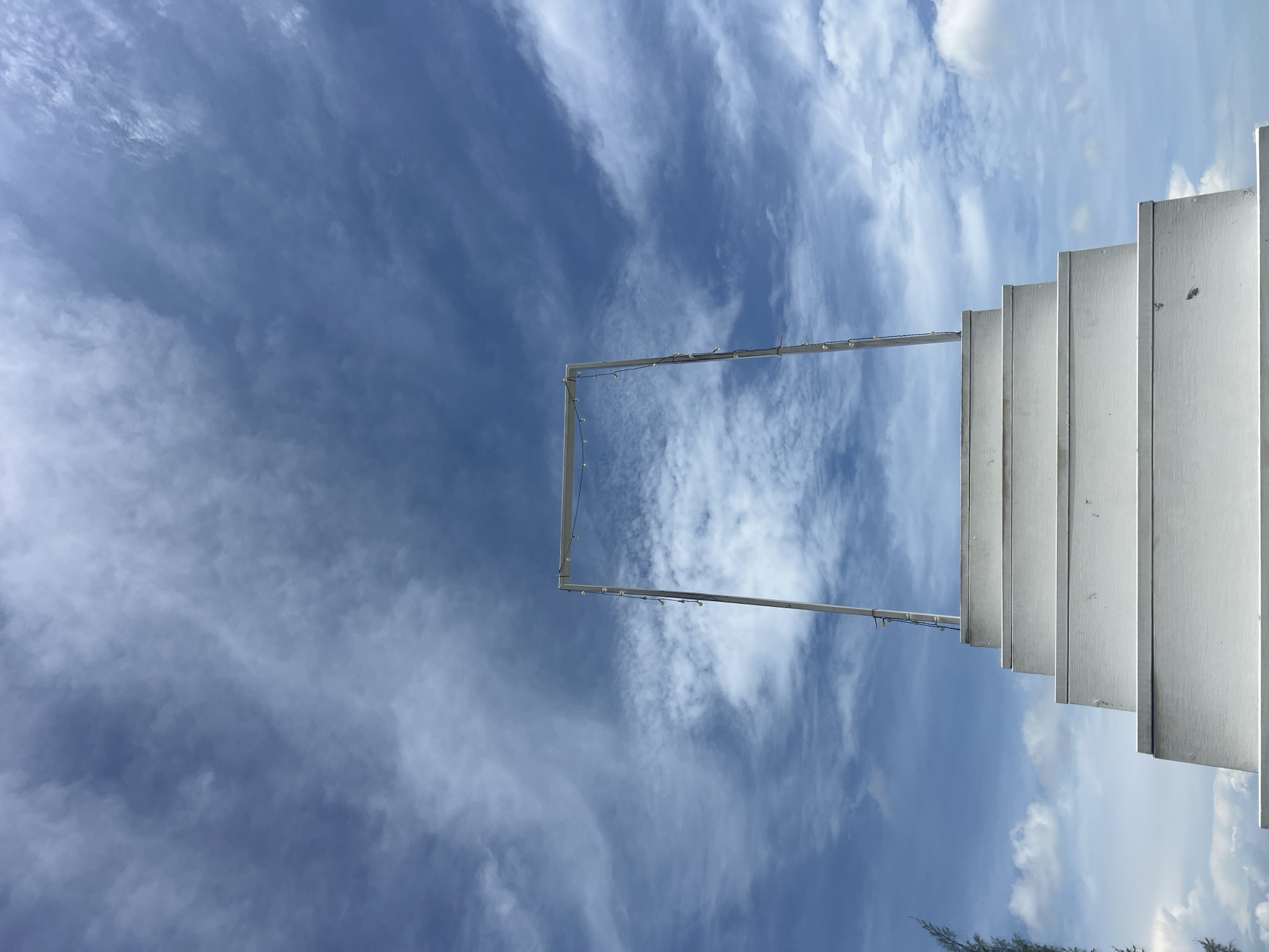 The image captures a stark, minimalist scene set against a bright blue sky filled with wispy white clouds. In the foreground, a set of clean, white wooden stairs ascends towards the sky. The stairs are evenly spaced and appear to be made of planks. At the top of the stairs, a simple, rectangular frame made of light-colored metal stands upright. This frame is adorned with a string of small, incandescent lights. The overall impression is one of a pathway leading to an open expanse, suggesting a sense of ascent or passage. The location is identified as Baan Thurian, Thailand, and the environment appears to be outdoors, possibly a coastal or elevated area given the vastness of the sky and the sparse greenery visible on the far left. There are no people or animals present in the image.