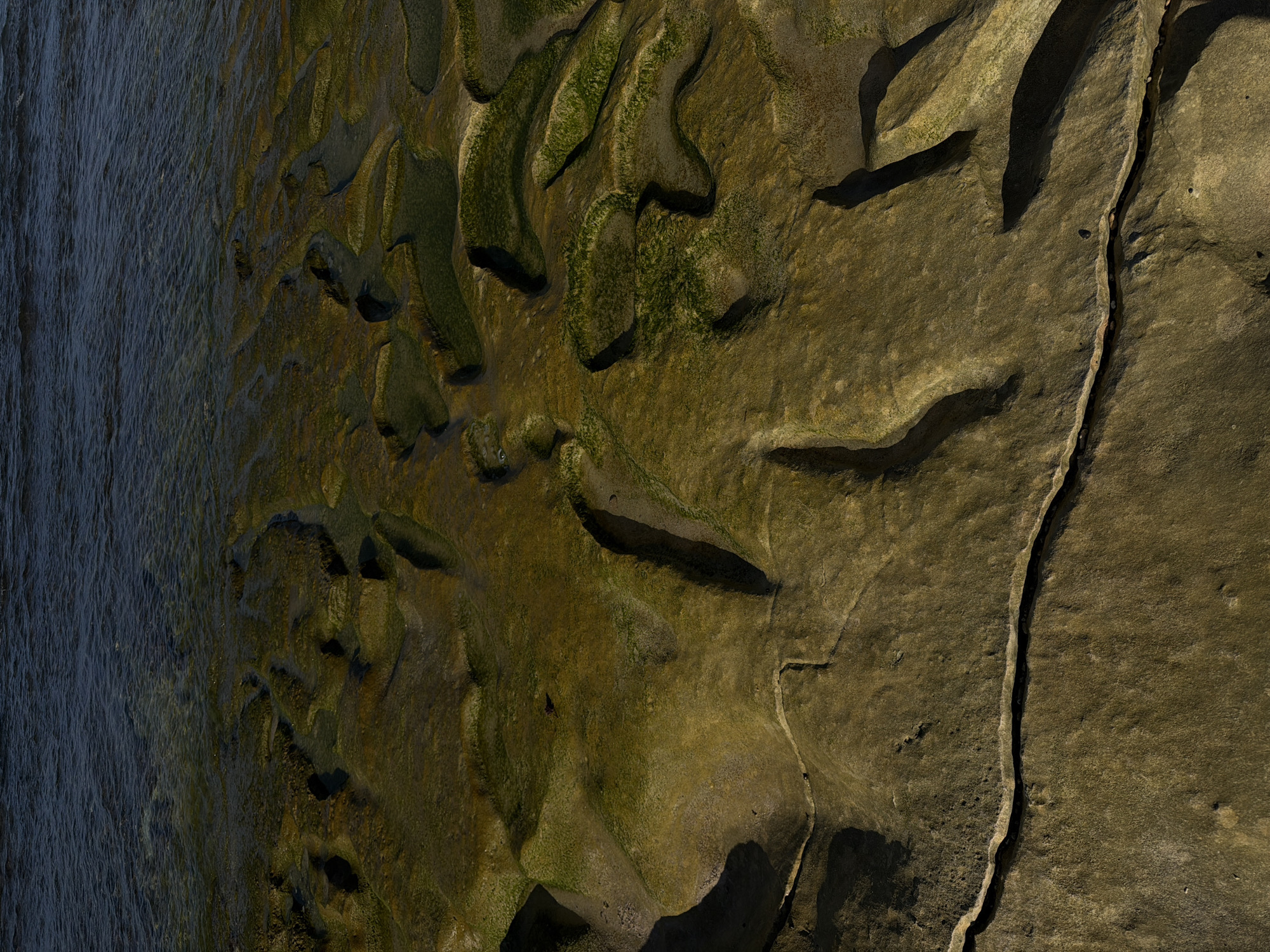 This aerial image depicts a landscape where land meets water. On the left is a body of water, potentially the ocean, with subtle wave patterns. The landmass transitions from a wet, moss-covered area to a drier, rocky terrain on the right. A prominent crevice runs down the right side of the image. The lighting suggests it is either sunrise or sunset. The scene appears uninhabited by people, concentrating on natural geological features in Kuta Selatan, Indonesia.