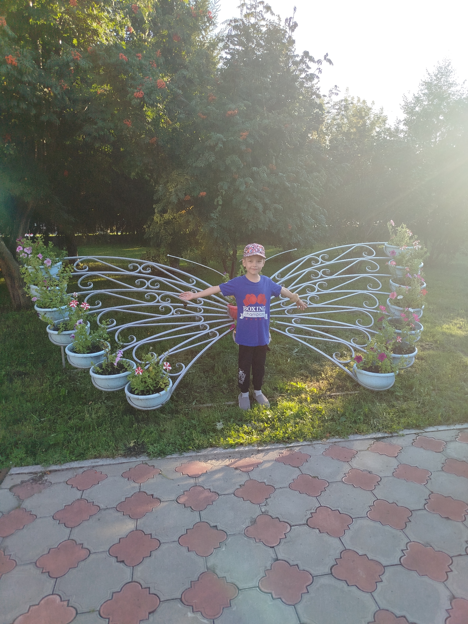 A young person wearing a blue t-shirt with a "BOXING champion" graphic and a patterned baseball cap stands with arms outstretched in front of a decorative white metal structure shaped like butterfly wings. The structure is adorned with multiple pots of pink and white petunias. The scene is outdoors, with a grassy area in the foreground leading to a patterned paved walkway. Behind the structure, there are trees with visible orange berries, suggesting it might be autumn. The sunlight creates lens flare in the upper right portion of the image. The location is identified as Yalutorovsk, Russia.