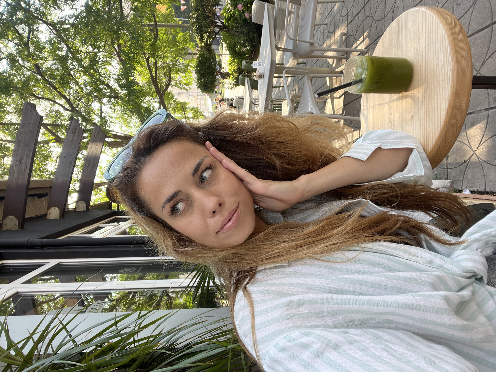 A woman with long, blonde hair is seated at a round wooden table outside a cafe in Bar, Montenegro. She is wearing a striped shirt and resting her hand on her face, looking to the side. On the table in front of her is a glass of green liquid with a straw. In the background, there are outdoor cafe tables and chairs, as well as trees and foliage. The sunlight suggests it is daytime.