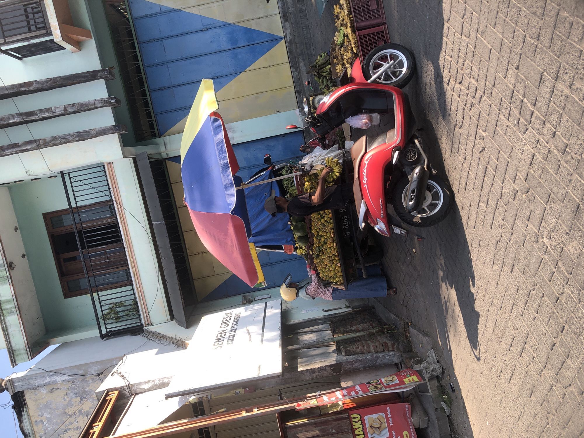 In the city of Kludan, Indonesia, a vibrant street scene unfolds beneath a clear sky. A woman in a yellow helmet and blue skirt, with a pink patterned shirt, stands by a cart laden with bright yellow bananas. Beside her, a man wearing a grey cap and black t-shirt sorts through another bunch of bananas. A red scooter, emblazoned with the word "Scoopy," is parked in front of the fruit stand, its sleek design contrasting with the rustic charm of the scene.
The fruit stand is shaded by a colorful umbrella, a cheerful canopy of red, blue, and yellow stripes. Crates overflowing with oranges and more bananas are stacked around the stand, suggesting a bountiful harvest. Behind the fruit stand, a building with a light blue facade and dark blue shutters stands tall, its architecture reflecting the local style. A sign on the building reads "SEMEN GRESIK KOKOH TAK TERBANDUNG," indicating a business nearby.
The street itself is paved with textured bricks, worn smooth by the passage of time and countless footsteps. The sunlight casts long shadows, creating a dynamic interplay of light and dark across the scene. To the left of the fruit stand, a red sign displays images of food items, hinting at the culinary offerings of the area. The overall impression is one of lively commerce and everyday life in a bustling Indonesian town.