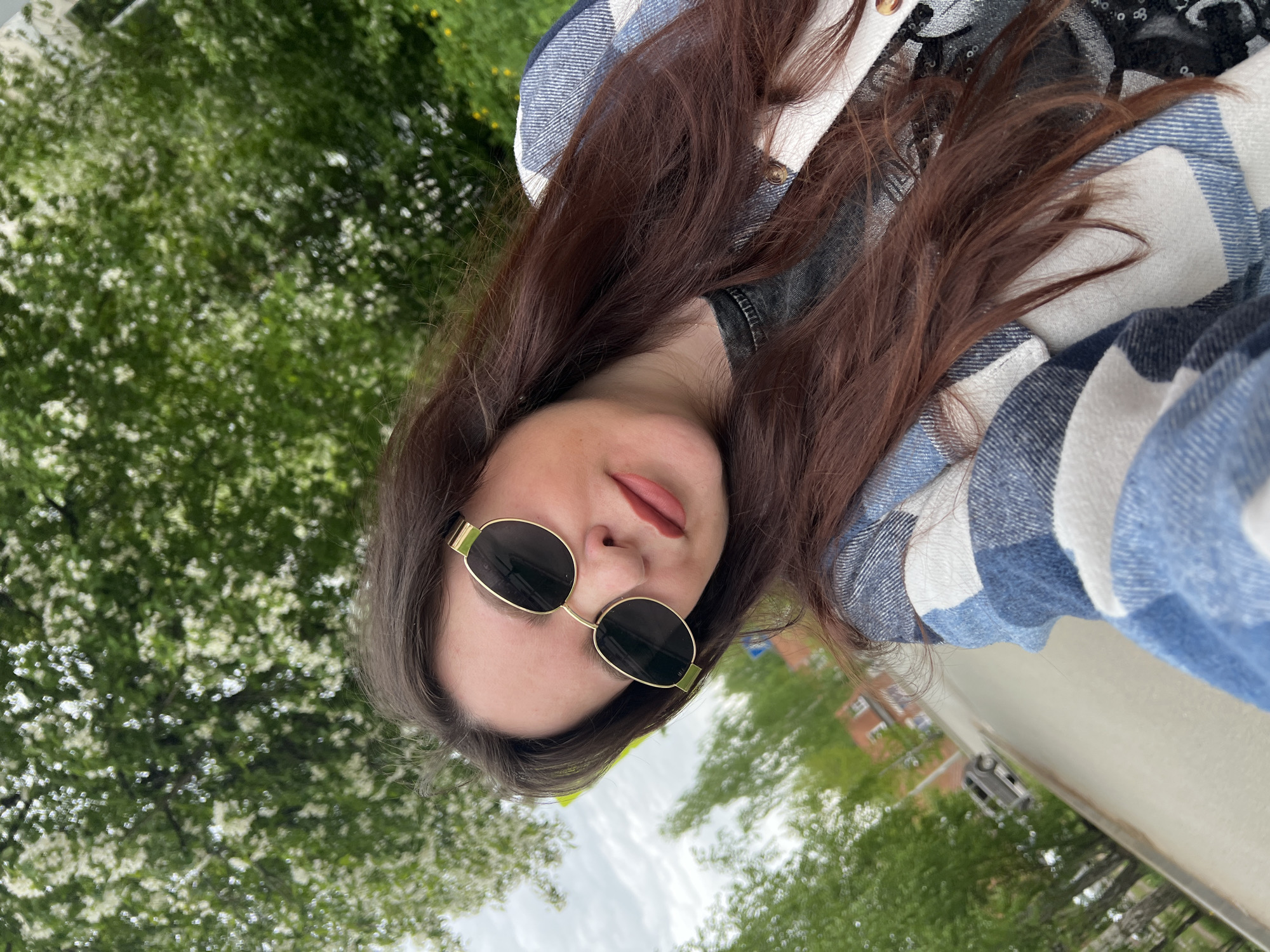 A woman with shoulder-length brown hair, wearing gold-rimmed sunglasses, a denim jacket, and a blue and white plaid flannel shirt is the subject of this outdoor selfie. She is sticking her tongue out slightly. The background consists of green trees and flowering white blossoms. A building and a paved street can be seen in the lower right. This location is Noviy, Russia.