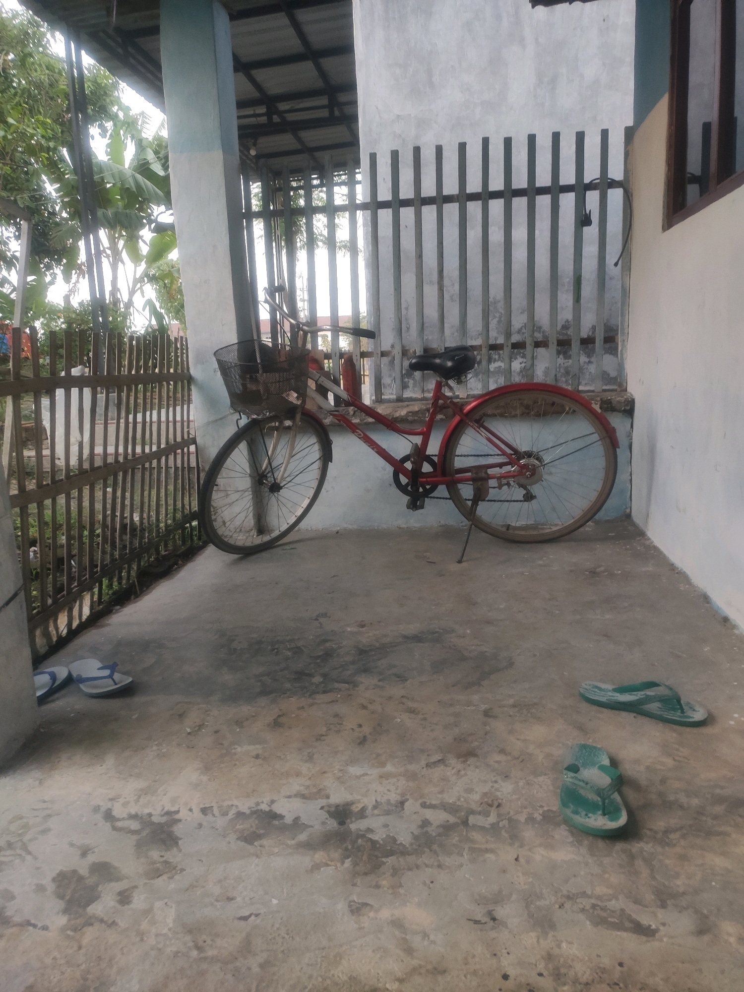 A red bicycle with a wire basket is parked on a concrete floor next to a metal fence and a white building. Two pairs of flip-flops, one blue and white and one green, are scattered on the floor. The scene appears to be an outdoor covered area, possibly a patio or carport, with foliage visible behind the fence. The lighting suggests it is daytime. The location is identified as Bongkot, Indonesia.