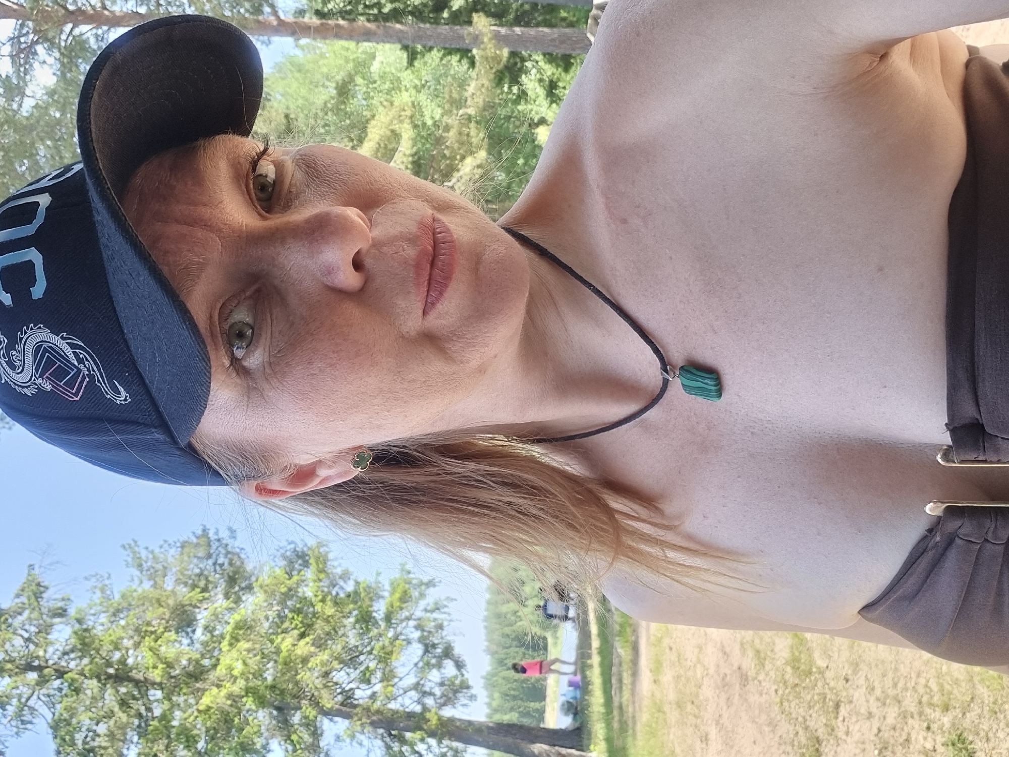 A woman is centered in the image, wearing a dark baseball cap with white lettering and a dragon design, and a brown top. She has fair skin, freckles, and blond hair. Her jewelry includes a black cord necklace with a green pendant and a green clover-shaped earring. She is outdoors on a bright day, with trees visible in the background. A body of water and several people are visible in the distance. The location cannot be determined by the image alone.