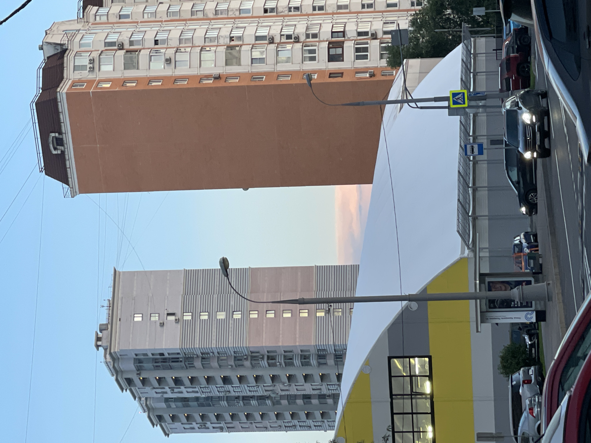 The image depicts a street scene in Moscow, Russia, likely in a residential or mixed-use area. Two tall apartment buildings dominate the background. The building on the left is lighter in color with horizontal grey and pink stripes and numerous windows and balconies. The building on the right is a taller, reddish-brown brick structure with a dark mansard roof and lighter colored apartments visible on its side. In the foreground, a large, modern building with a white, arched roof and a yellow band runs horizontally across its facade. This building appears to be a sports complex or similar facility. A street lamp stands tall in the middle of the scene, and a bus stop shelter with advertisements is visible near the lower right. Several cars are present on the road, including a dark SUV in the foreground and other vehicles parked or driving in the background. The sky is a pale blue with some light clouds visible. The lighting suggests it is either early morning or late evening. The overall impression is a contemporary urban environment.