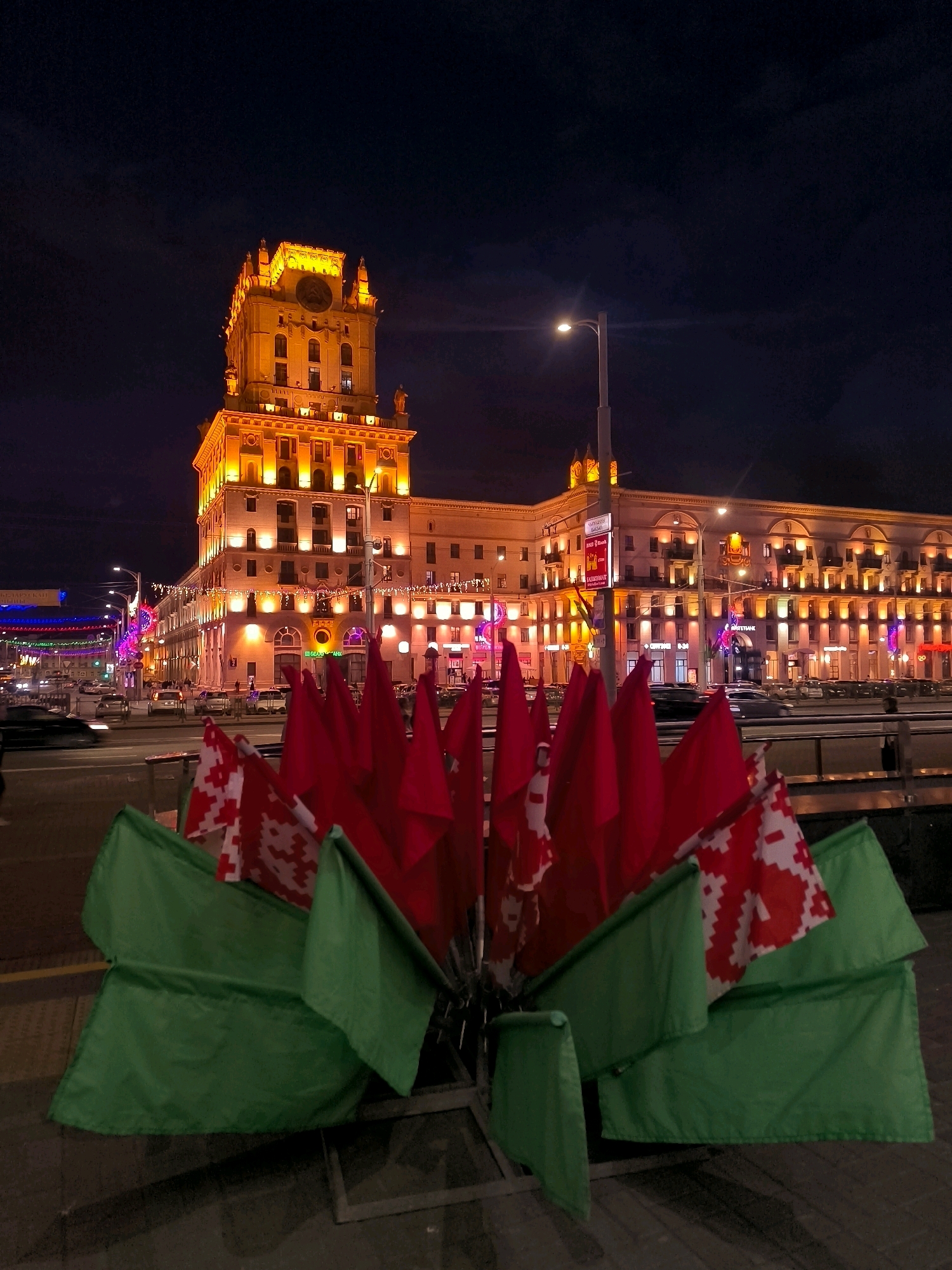 At night in Minsk, Belarus, a large, illuminated building stands prominently. The building is multi-storied with a prominent tower featuring a clock. It is lit with warm, orange lights, highlighting its architectural details. In the foreground, a display of red and green flags is arranged in a fan-like pattern. Some of the red flags have a white and red pattern, resembling the historical flag of Belarus. The flags are on a metal frame, and some green flags are draped around the base. The scene is set on a street with traffic visible, including cars in motion. Streetlights and building lights create a vibrant atmosphere. The sky is dark, indicating nighttime. The overall impression is of a city illuminated at night, with a patriotic display in the foreground.