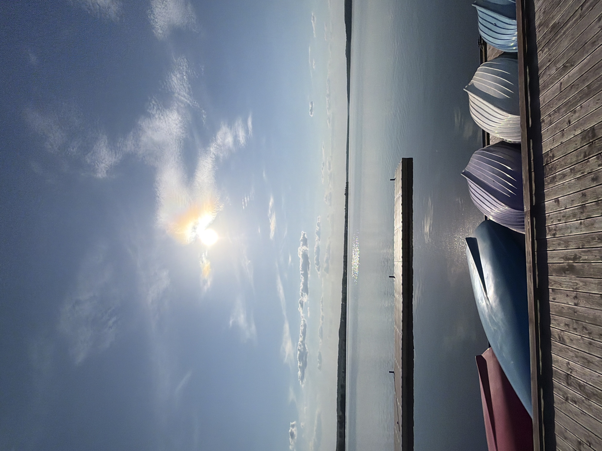 The image depicts a lakeside scene in Votkinsk, Russia, under a bright sun and partly cloudy sky. A wooden dock extends into a calm body of water, likely a lake or reservoir. Several small boats are neatly stacked along the dock's edge, exhibiting various colors: blue, white, and purple. The sun's glare reflects on the water's surface, creating a sparkling effect. The opposite shore is visible in the distance, marked by a thin line of land. The focus is on the natural environment and recreational boating.