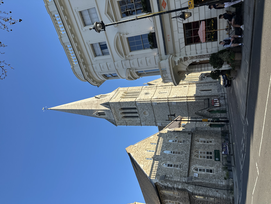 An outdoor scene captured under a clear blue sky depicts the intersection of Pimlico Road in Pimlico, London SW1, featuring St Barnabas Church and The Orange pub. On the left, St Barnabas Church is a prominent Gothic Revival structure constructed of light-colored, rough-hewn stone, featuring a tall, slender spire topped with a weather vane. Its facade displays multiple pointed arch windows, and a sign identifying "St Barnabas" is visible on the lower section of an adjacent church building. To the right, The Orange pub is a white, curved building exhibiting classical architectural elements, including decorative cornices and a balustrade. The pub's ground floor features large windows, an awning, and signage for "PUB" and "The Orange." A dark street lamp post with two lanterns stands immediately in front of the pub. On the paved sidewalk in front of The Orange, three adult males are present: two are seated at a small outdoor table under a red and white striped umbrella, and one stands nearby to their right. The asphalt road in the foreground, marked with white lines, curves to the left. Sunlight casts distinct shadows across the scene, particularly on the church facade and the road. A dark-colored vehicle is partially visible parked on the street near the church. Bare tree branches are visible against the sky in the upper right quadrant.
