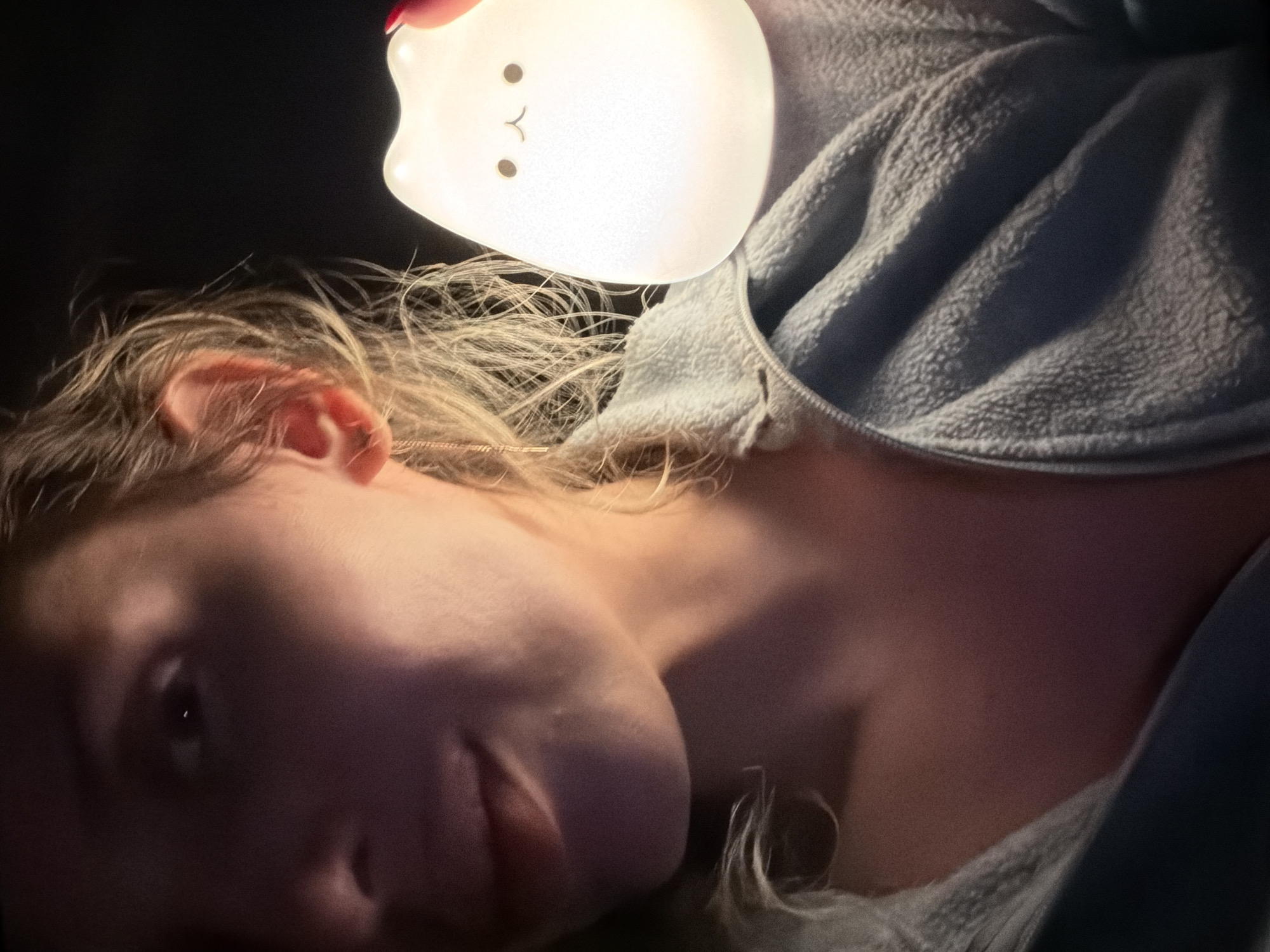 An intimate, low-light portrait shows a person, likely a young woman, lying down in what appears to be a relaxed indoor setting in Budva, Montenegro. Her head is positioned to the left, with her face partially visible and illuminated by a glowing, white cat-shaped lamp held above her head, casting a soft, warm light. Her fair skin and light-colored, wavy hair are highlighted by this light, while much of her face remains in shadow, particularly her right eye and the lower half of her face.
The person is wearing a light-colored, possibly grey or light blue, fuzzy or fleece garment, resembling a hoodie or jacket, which is partially visible on the right side of the frame. A faint golden chain or string is visible near her left ear. A fingertip with red nail polish can be seen at the top, holding the cat lamp. The lamp itself has simple black dots for eyes and an upside-down 'v' for a mouth, giving it a friendly, minimalistic expression. The background is predominantly dark, indicating that the photo was taken at night or in a dimly lit room, with the lamp serving as the primary light source. The overall mood is peaceful and calm, suggesting a quiet moment of relaxation. No visible text is present in the image.