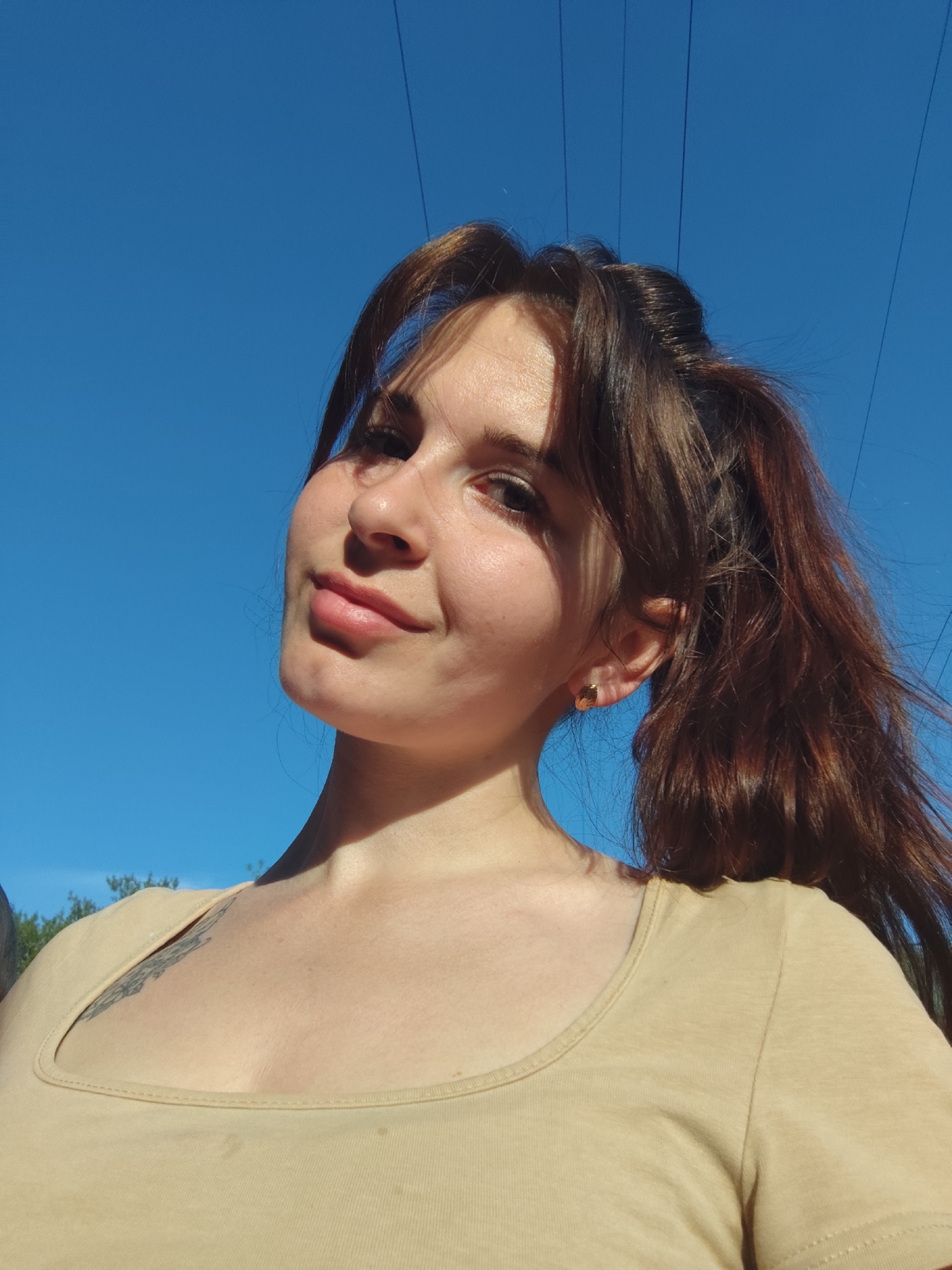 A young woman with light skin, dark brown hair pulled back into a ponytail, and a beige shirt is looking at the camera. Her hair has bangs, and she is wearing gold earrings. She has a tattoo on her upper chest. The background is a bright blue sky. Power lines are visible above her head. The location is Фоки, Russia.