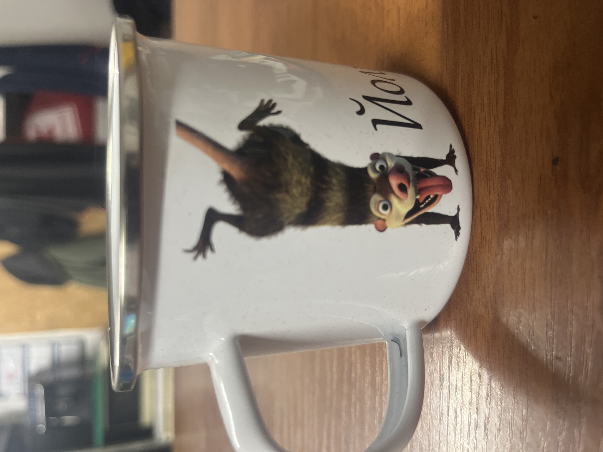 A white mug with a metal rim sits on a wooden table. The mug features an image of a character from the movie Ice Age, an opossum with its head upside down and tongue out, and Cyrillic lettering that appears to say "Йоли" (Yoli). The background is blurred and suggests an indoor environment with some shelving and dark clothing hanging. The mug is the primary subject, positioned centrally in the frame. The lighting suggests an indoor setting, possibly a home or office. There are no people or animals present in the immediate scene. The location is not specified in the image, nor is it possible to determine it from the visual information.