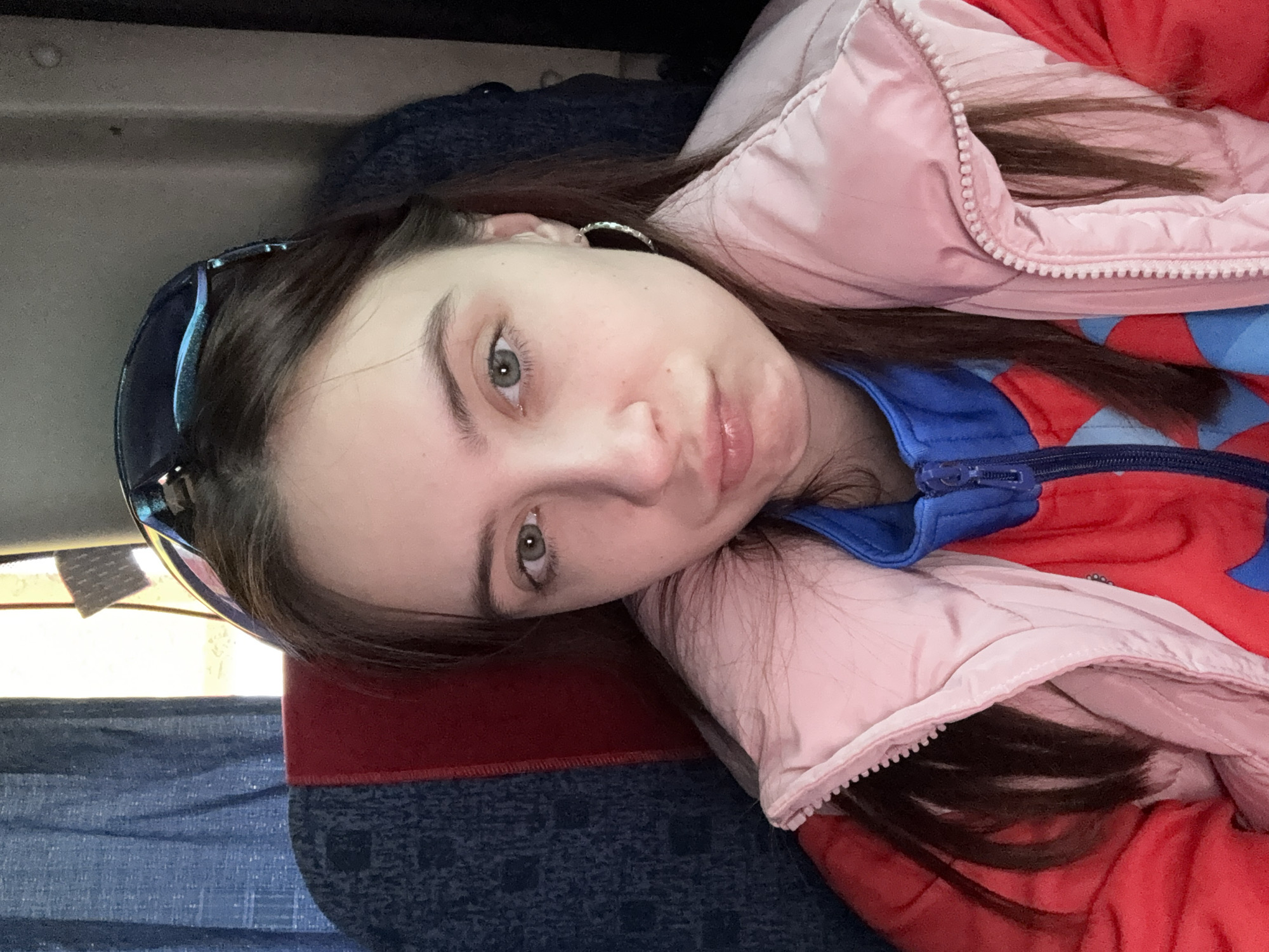 A young woman with fair skin, brown hair, and green eyes is positioned lying down with her head tilted slightly to the left. She is wearing sunglasses perched on her head, a silver earring, and a colorful jacket with pink, red, and blue sections. The background appears to be the interior of a vehicle or airplane, with a seat visible behind her that is patterned with blue and red.
There is no data on this image to support this as being in the city of Челва, Russia.