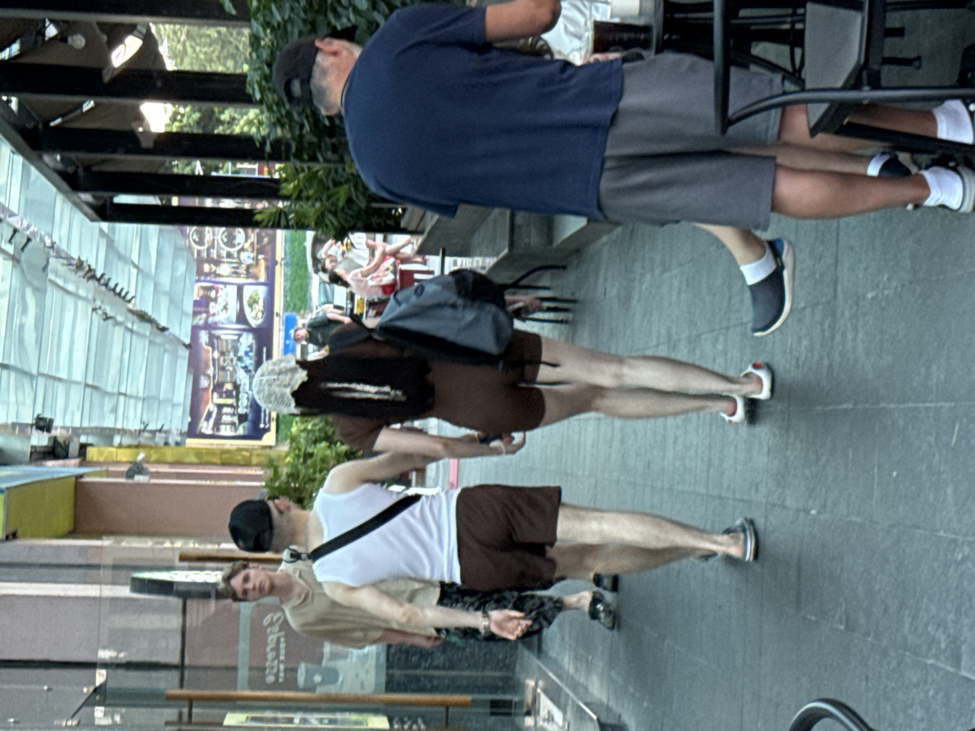 The image shows a street scene in Nha Trang Ward, Vietnam, with several people walking on a tiled sidewalk. In the foreground, a man wearing a blue t-shirt and grey shorts is walking away from the camera, with his back to the viewer. He is wearing black sneakers with white socks. To his left, a woman in a brown outfit and a white head covering is walking away from the camera, holding hands with a man in a white tank top and brown shorts. The man is wearing black sandals. Another man in a light-colored shirt and patterned shorts is walking away from the camera to the left of the woman and man. In the background, there are shops with awnings and posters, and trees. The lighting suggests it is daytime.