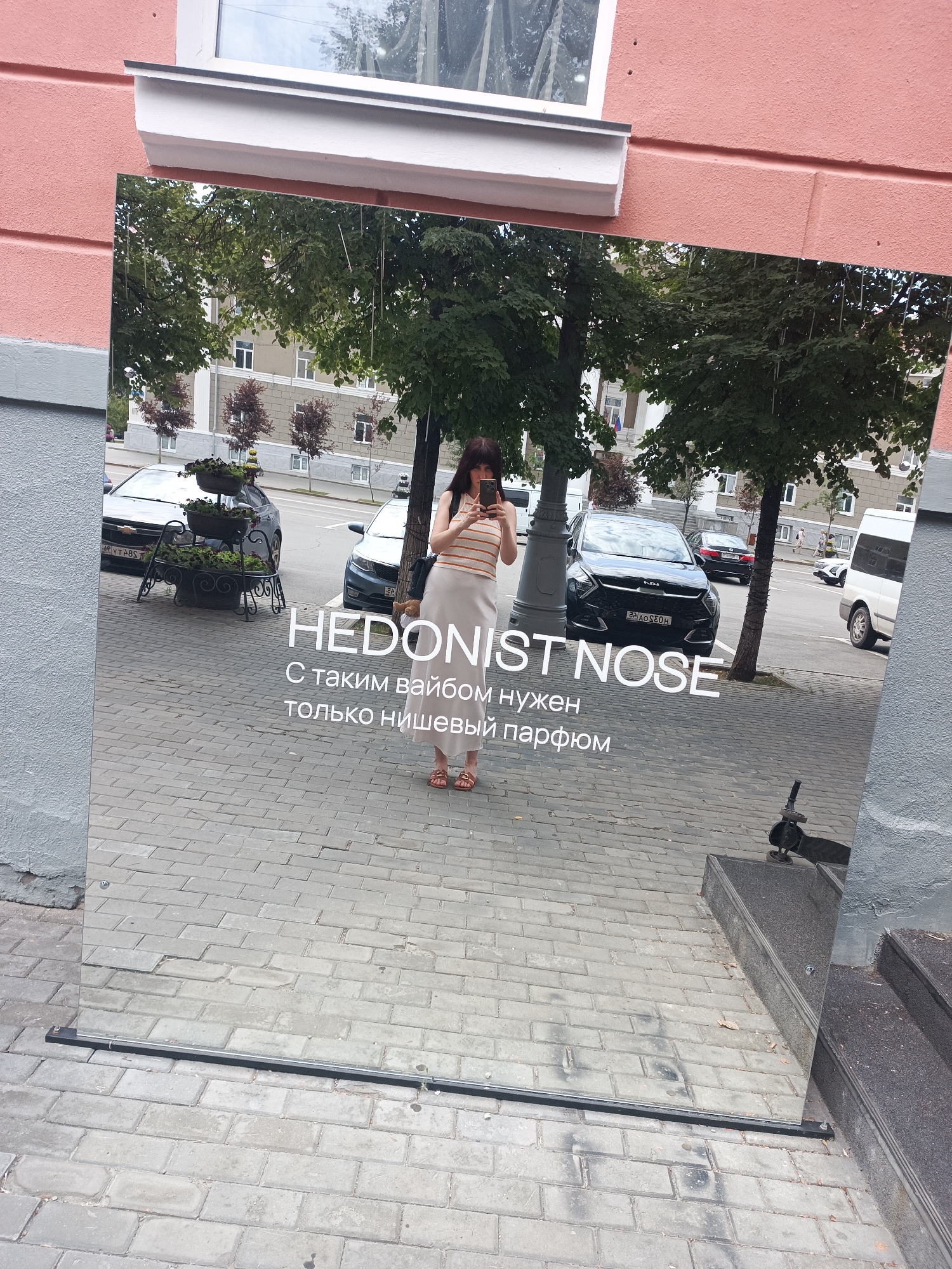 In Kurgan, Russia, a full-length mirrored sign stands on a sidewalk made of gray cobblestones, leaning against a pink-colored building. The mirror reflects a street scene with trees, cars, and buildings, as well as a woman posing for a selfie. The woman, wearing a striped shirt and a light-colored skirt, is holding a smartphone and looking at the screen. She is standing on the cobblestone sidewalk, partially reflected in the mirrored sign.
Text is visible on the mirrored sign, reading "HEDONIST NOSE" in large white letters. Below this, smaller white text in Russian states, "С таким вайбом нужен только нишевый парфюм," which translates to "With such a vibe, only niche perfume is needed."
Several cars are reflected in the mirror, including a black sedan with a license plate that appears to be "29 AO 520 H" and a white van. In the background, trees with green foliage are visible, along with buildings that have multiple windows. The environment appears to be an urban setting during the daytime.