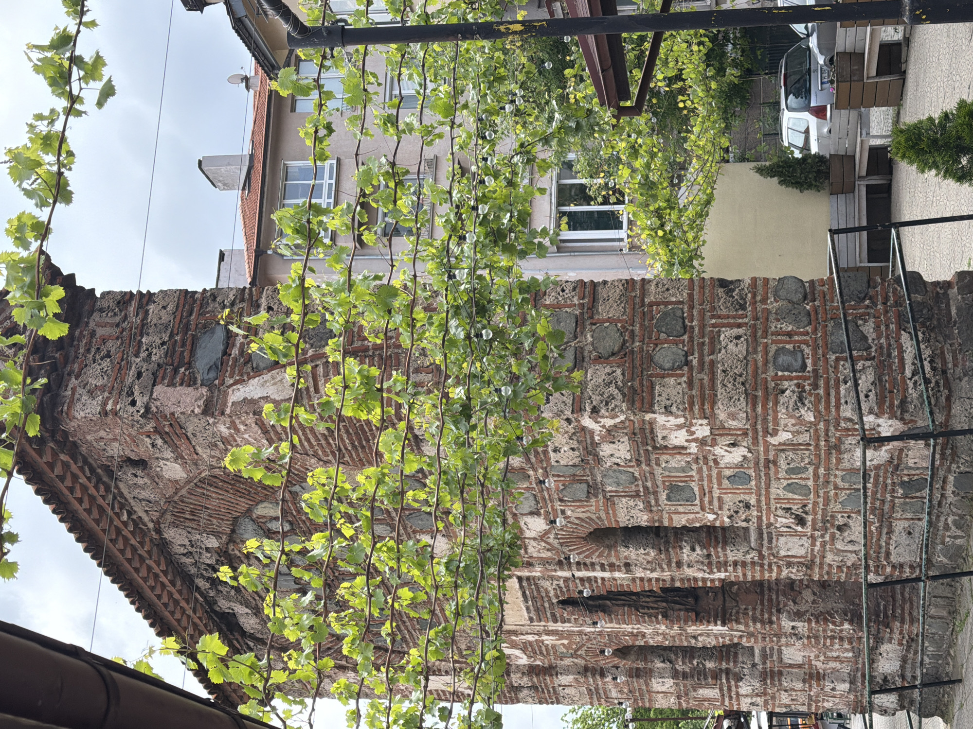 The image shows the Rotunda of Saint George in Sofia, Bulgaria. The architectural structure is made of red bricks and stones, decorated with green vine plants hanging from above. In the background, a light-colored building with windows is visible, along with a white car and some greenery. The weather appears to be overcast.