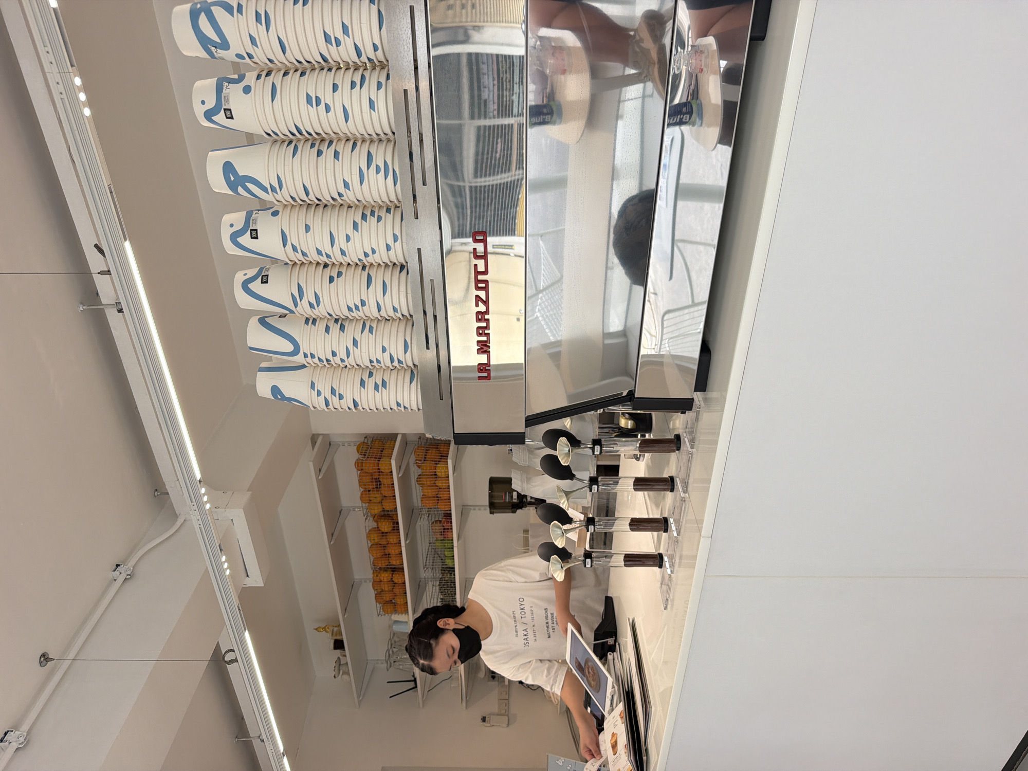 In a brightly lit cafe interior, a young woman wearing a black face mask and a white t-shirt stands behind a polished chrome counter. The t-shirt has text that includes "OSAKA / TOKYO" and "MAYHEM VISIONS 1ST AVENUE". She is holding a tablet and looking at a menu or order sheet. In front of her, on the counter, are four glass tubes containing what appears to be coffee grounds, each topped with a scoop. A large, professional espresso machine with "LA MARZOCCO" branding is prominently displayed to the right of the woman. Above the counter, stacks of disposable paper cups with blue wave-like designs are neatly arranged. Shelves behind the woman hold baskets filled with oranges and some limes. The environment is clean and modern, with white walls and ceiling, and fluorescent lighting. The reflections in the espresso machine suggest the presence of tables and chairs, and possibly other patrons, in the cafe. This cafe is located in Bangkok, Thailand, as indicated by the text on the woman's t-shirt.