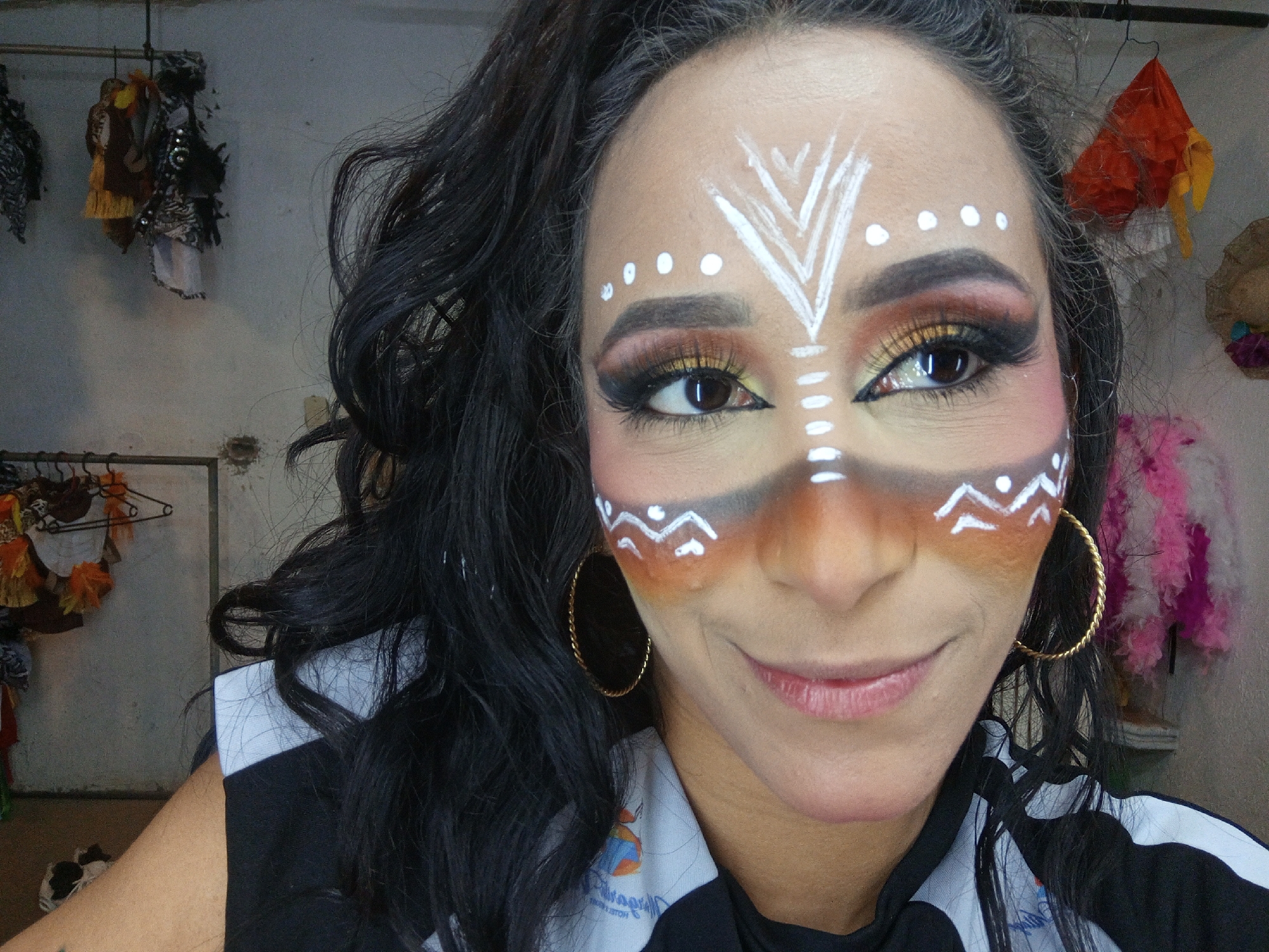 A close-up selfie shows a woman with elaborate face paint and dramatic makeup, smiling gently as she looks slightly to the right of the camera. Her face is adorned with white geometric patterns, including chevrons on her forehead, dots above her eyebrows, vertical lines extending from below her eyes, and wavy lines on her cheeks. Her eye makeup features striking gold and brown eyeshadow, heavy winged eyeliner, and long false eyelashes, with a bright yellow accent on her lower waterline. Brown, orange, and black paint is blended around her eyes and nose, creating a contoured effect.
She wears large gold hoop earrings and has long, dark, wavy hair that falls over her shoulders. Her attire includes a black and white collared shirt, possibly a polo or jacket, with a partially visible blue and orange logo and some faint, reversed text on the fabric.
The background, a plain, light-colored wall, suggests an indoor setting like a dressing room, backstage area, or workshop. Various items are visible in the background: clothes or fabric decorations hang on a rod to the left, an orange fabric decoration hangs on the right, and a small straw hat-like item is partially seen to the far right. The scene appears well-lit, highlighting the intricate details of her makeup. This vibrant presentation suggests she is prepared for a performance, cultural event, or festival in Porlamar, Venezuela.