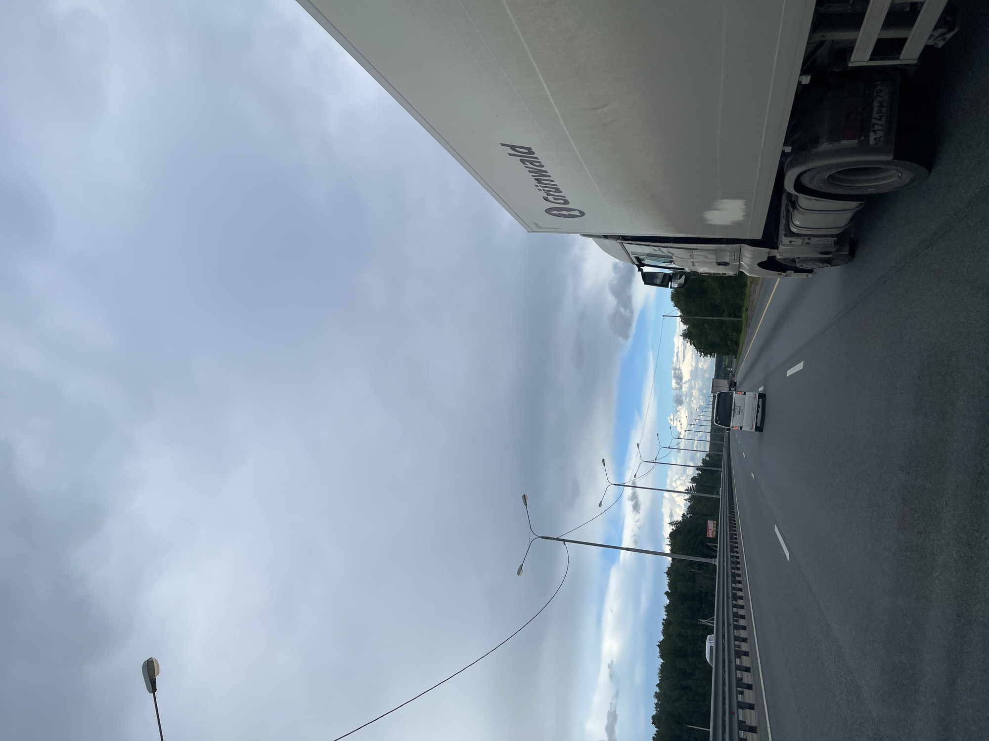 A daytime roadside view in Бавыкино, Russia, displays a multi-lane highway under an overcast sky. A white Grünwald truck is visible on the right, partially obscuring the view. Other vehicles are traveling in the distance. Streetlights line the highway. The shoulder of the road is visible to the left.