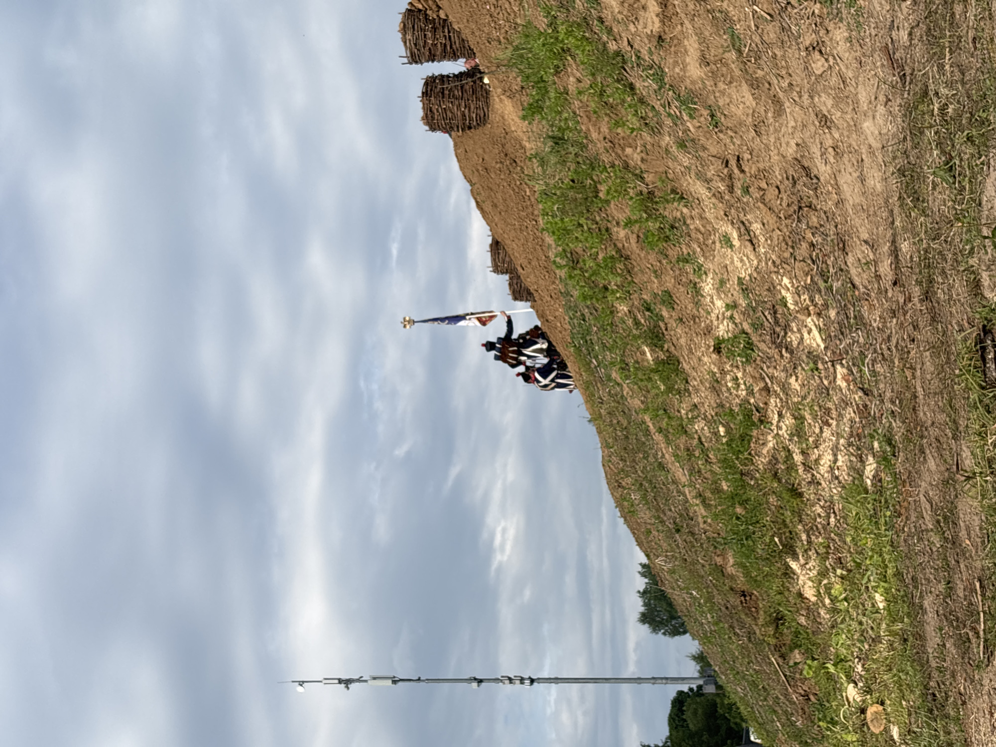 The image captures a historical reenactment event on a grassy, earthen embankment. In the background, a tall, modern cell tower stands against a cloudy sky. On the crest of the embankment, several individuals dressed in 18th-century military attire are positioned. One person, holding a flag with a golden eagle emblem and blue and white stripes, appears to be leading the group. Another individual in a similar uniform is visible behind them. The embankment is fortified with circular, woven wooden structures, likely representing temporary defensive emplacements. The foreground shows sparse vegetation and dry soil. The scene is set in an outdoor environment, with trees visible in the far left background. This event is taking place in Goncharovo, Russia.