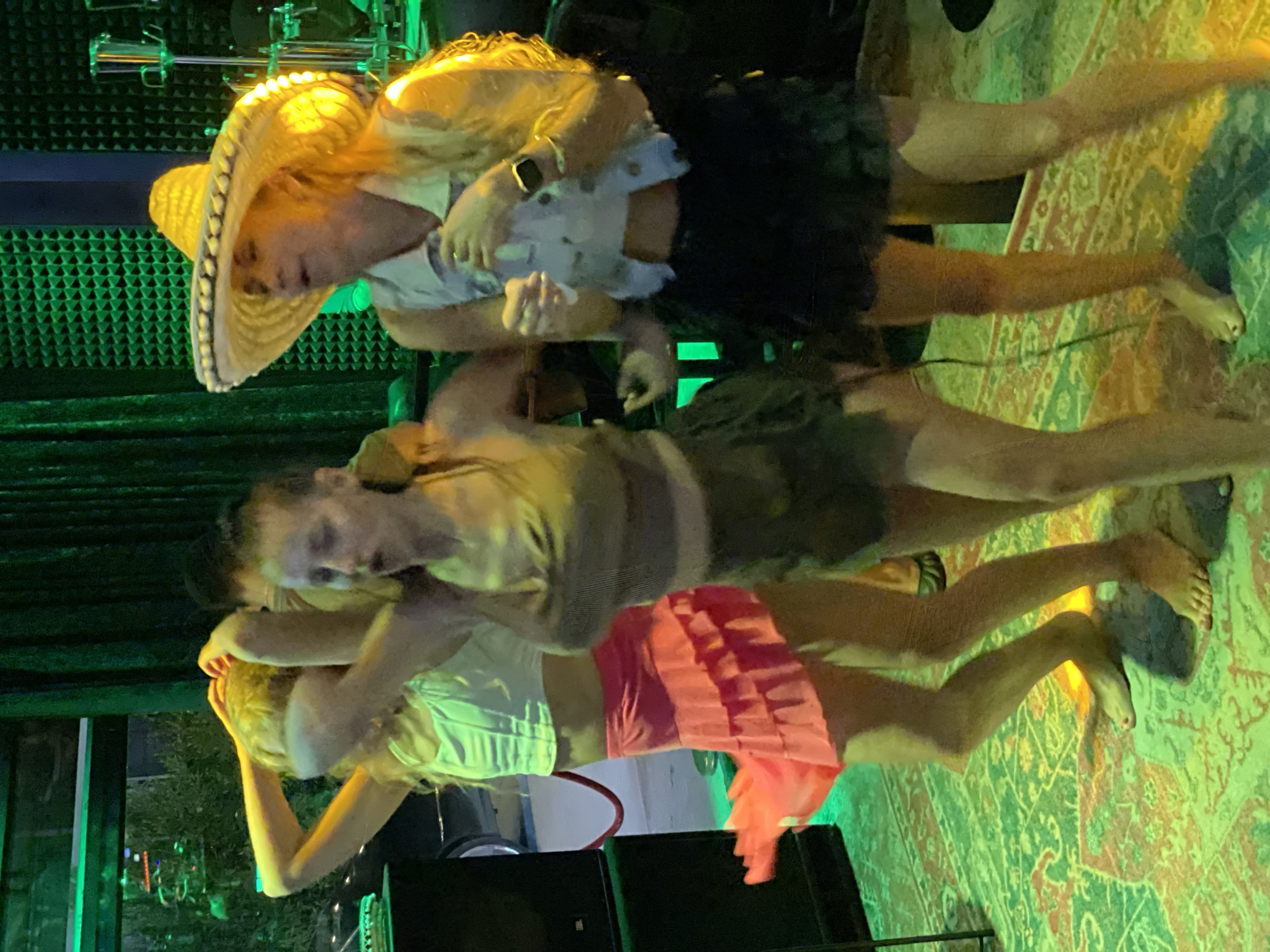 Three women are piled on top of each other, in a space with a green light wash. The top woman wears a sombrero, a light-colored collared shirt and a dark skirt, and is barefoot. The middle woman wears a tan top, and is also barefoot. The bottom woman wears a white top and a bright pink skirt. A rug is visible in the foreground. Behind the women, there is a green soundproof wall and musical instruments. The scene is at Baan Bang Rak, Thailand.