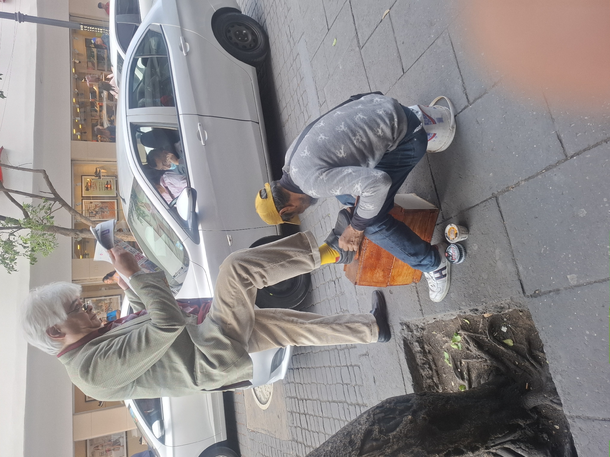 The image captures a street scene in Mexico City, Mexico. An older man, dressed in a blazer and corduroy pants, sits with one leg propped on a car tire, reading a newspaper. A younger man in jeans, a gray sweatshirt, and a yellow baseball cap is crouched near the older man's foot, seemingly working on or shining his shoe. A small, wooden box sits beside the younger man, along with a few cans. A white car is parked between them, and another person is visible inside the car. The background shows a storefront with display windows. The setting appears to be a sidewalk with a tree pit to the side.