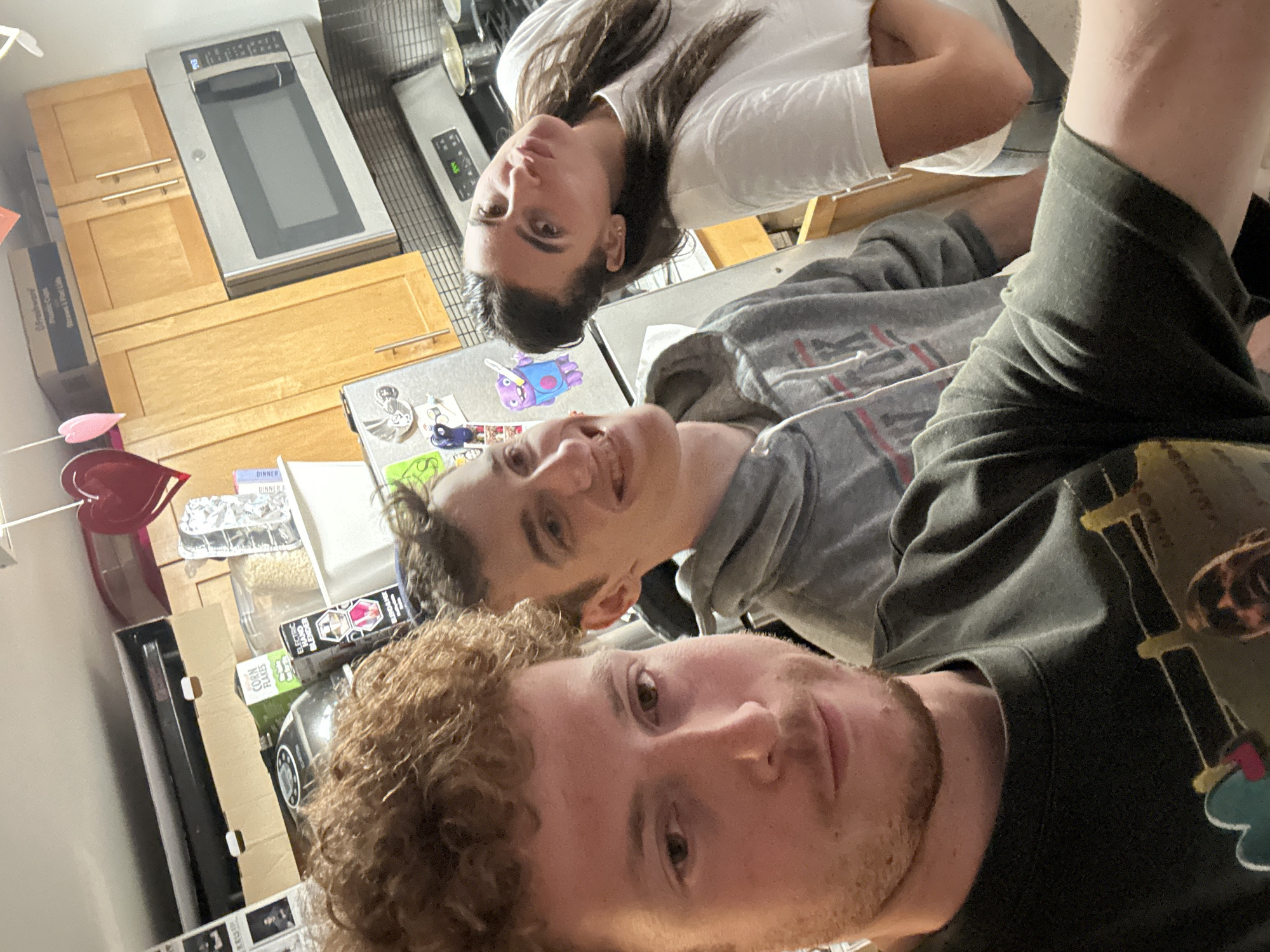 The image captures three individuals taking an informal group selfie in a home kitchen. The photograph is rotated 90 degrees clockwise, presenting the subjects and background at an unconventional, sideways angle.
Closest to the camera is a person with curly light brown hair and a light beard, wearing a dark green long-sleeved shirt. They look directly at the camera with a neutral expression. Behind them, a second person with short dark hair, wearing a grey hooded sweatshirt, is visible, smiling slightly and also looking towards the camera. Further back, and appearing upside down due to the image's orientation, is a third individual with long dark hair, wearing a white t-shirt, who is also looking at the camera.
The background shows a domestic kitchen setting. Light wooden cabinets are visible, above which a silver microwave oven is prominently placed, displaying "903" on its digital clock. To the right of the microwave, part of a stove or oven control panel shows "88.8". A refrigerator door, covered with various magnets including a purple character, is also visible. On the countertop, various items are scattered, including a red heart-shaped object, a silver baking tray with food (possibly lasagna), and product packaging. Visible text includes "CORN FLAKES," "ELECTRIC HAND BLENDER," "DINNER," and "Ofreshware Plastic Cups Straws & Flat Lids." The overall scene suggests a casual, relaxed moment among friends or family in an indoor environment within New York, United States of America.